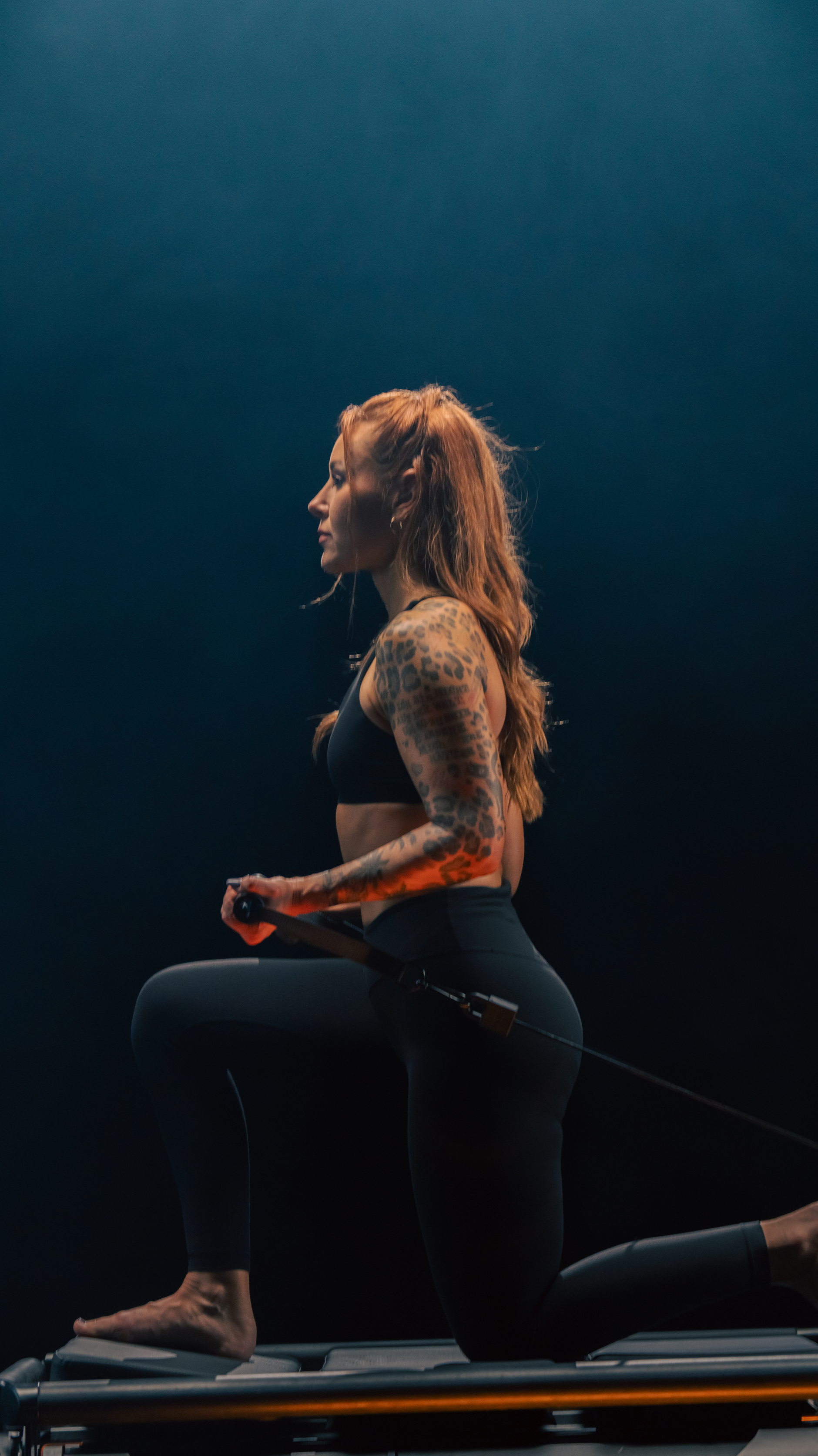 A woman with tattoos on her arms and long hair using a resistance band while kneeling on a Lagree EVO RAMP in a dark studio.