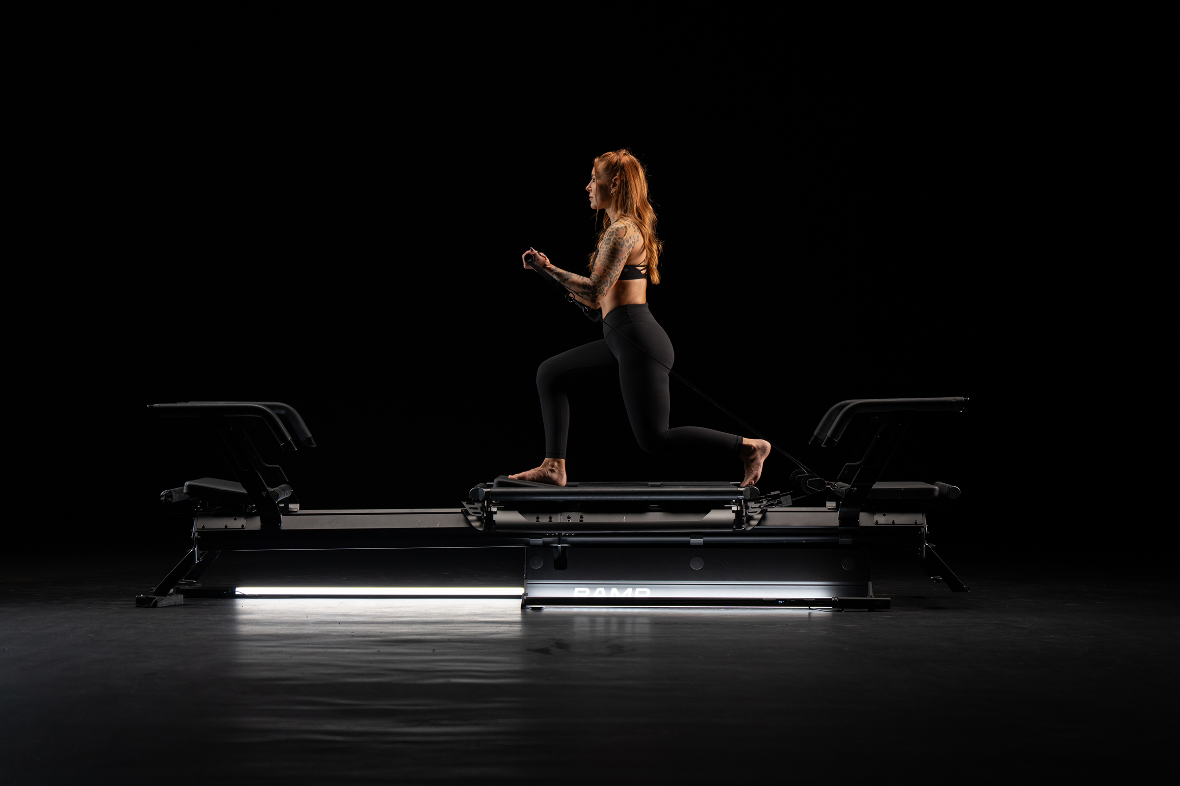A woman with red hair and tattoos wearing black workout clothes kneels on a Pilates reformer machine in a dark studio, illuminated by a single light underneath.