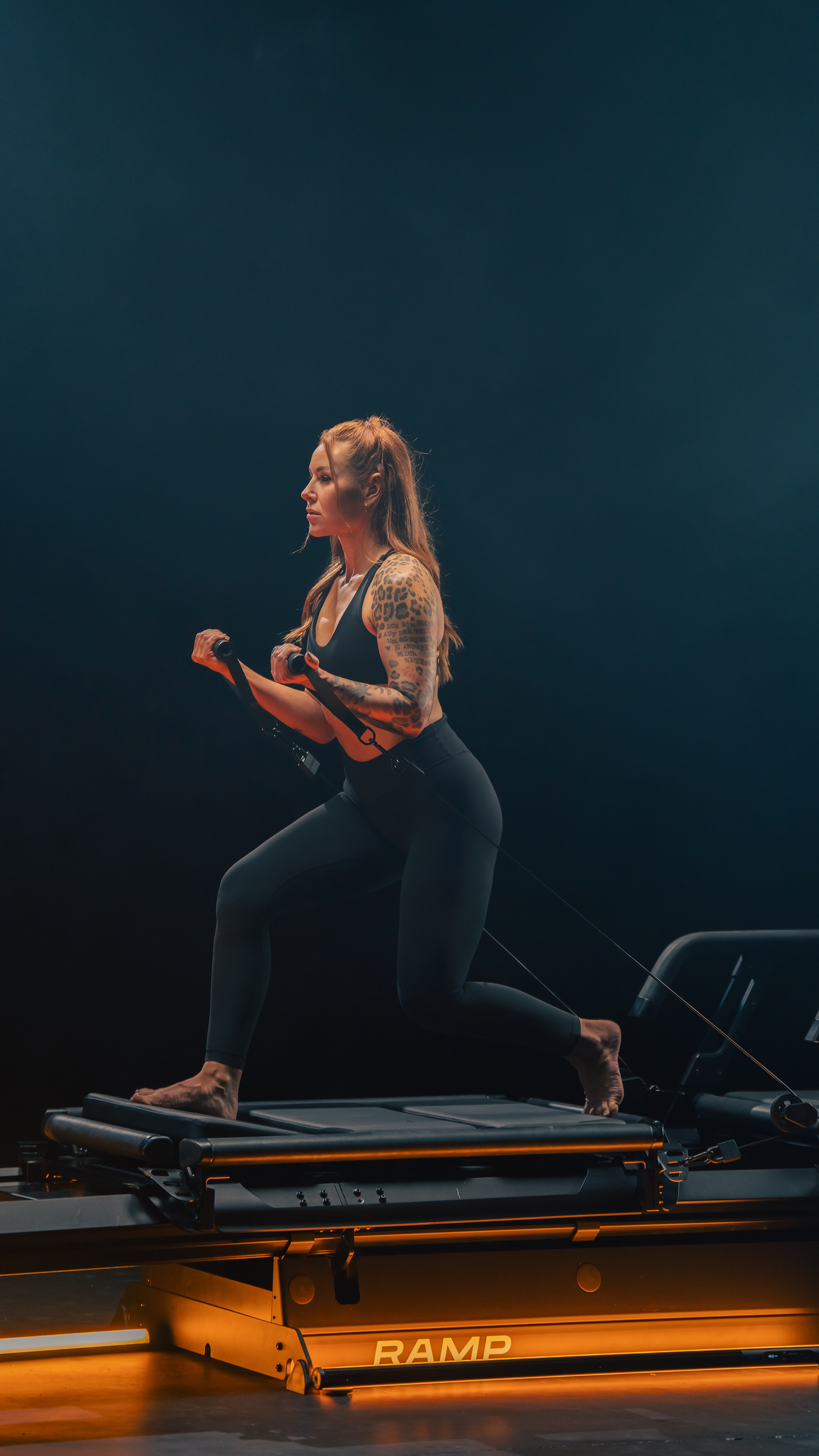 A woman exercising on a Lagree EVO RAMP with resistance bands in a dark gym setting.