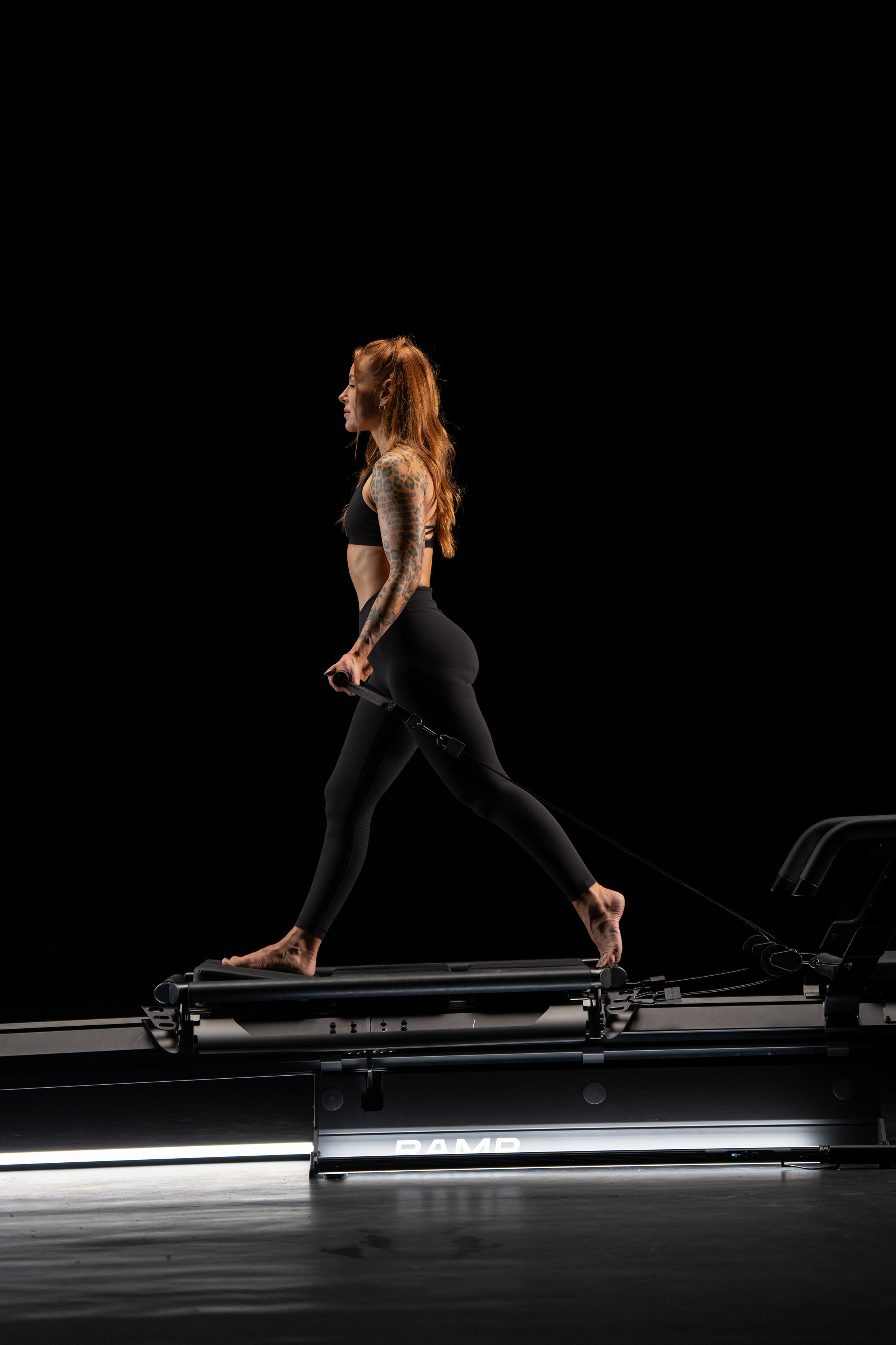 A woman with red hair, tattoos on her left arm, wearing black sportswear, is walking on a Lagree EVO RAMP in a dark studio.