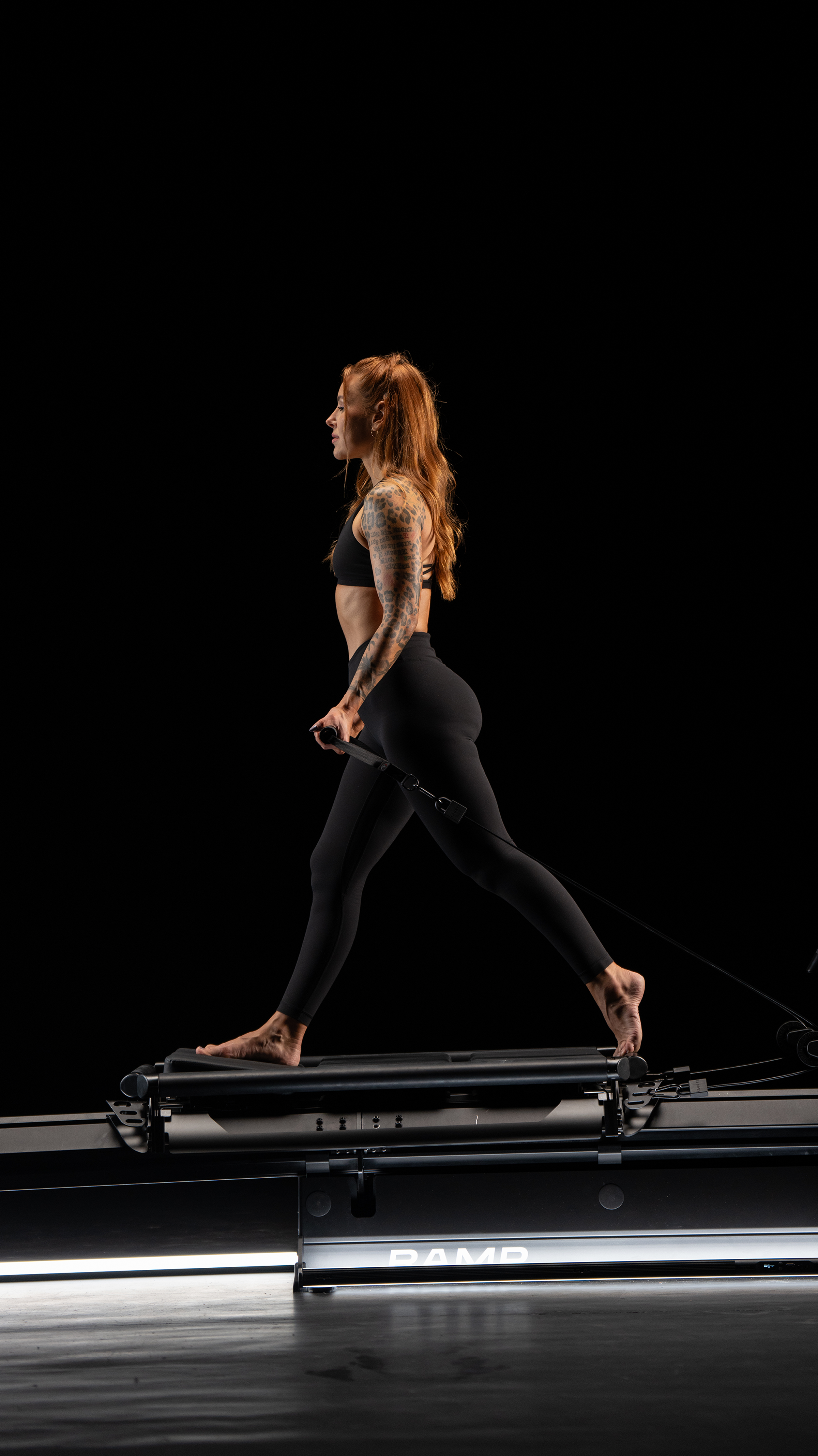 A woman with tattoos on her right arm performs a move on a Lagree EVO RAMP in a dark studio, wearing black athletic clothing.