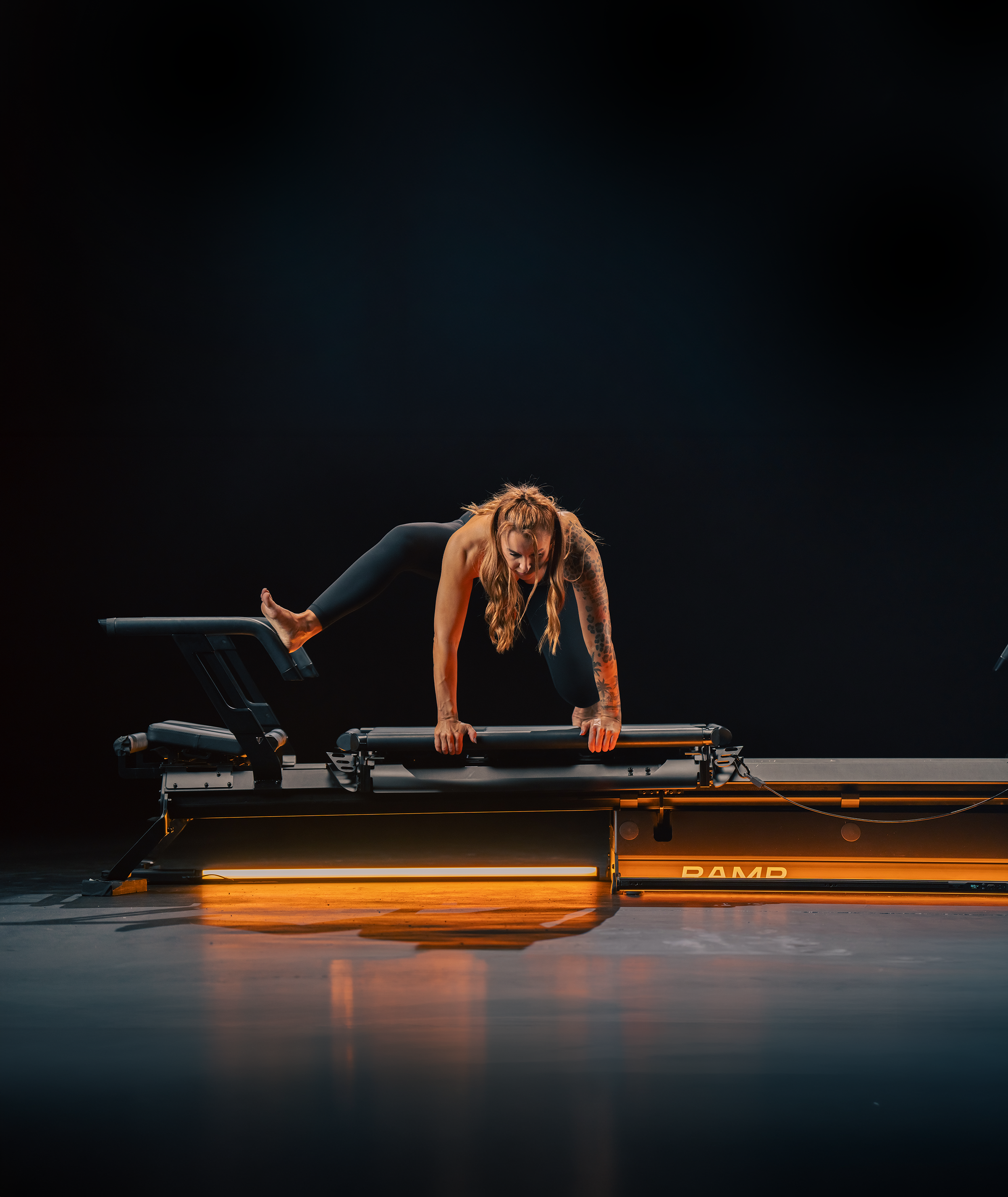 A woman with long red hair and tattoos on her arm, dressed in black workout clothing, is performing a yoga pose on a reformer machine in a dark studio with orange lighting.