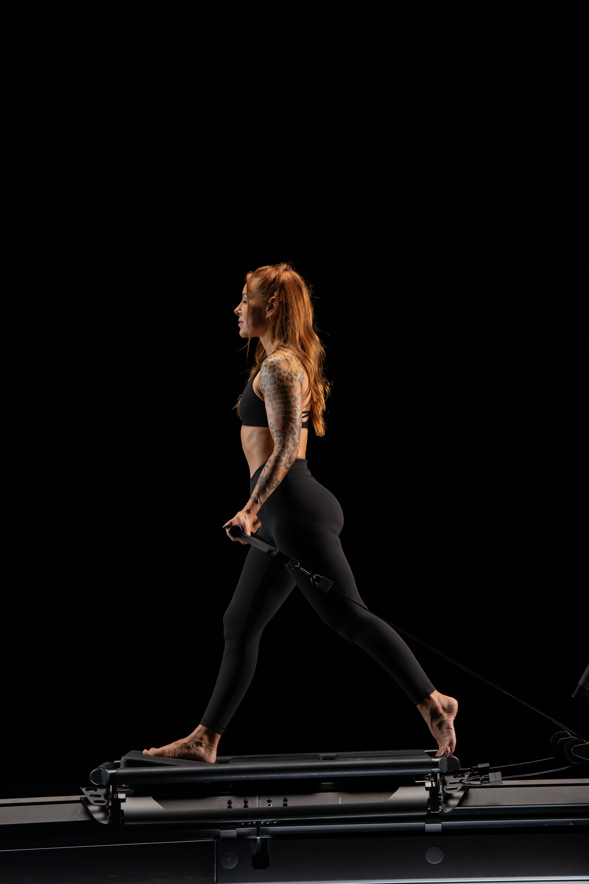 A woman with red hair, tattoos on her left arm, wearing black sportswear, is walking on a Lagree EVO RAMP in a dark studio.