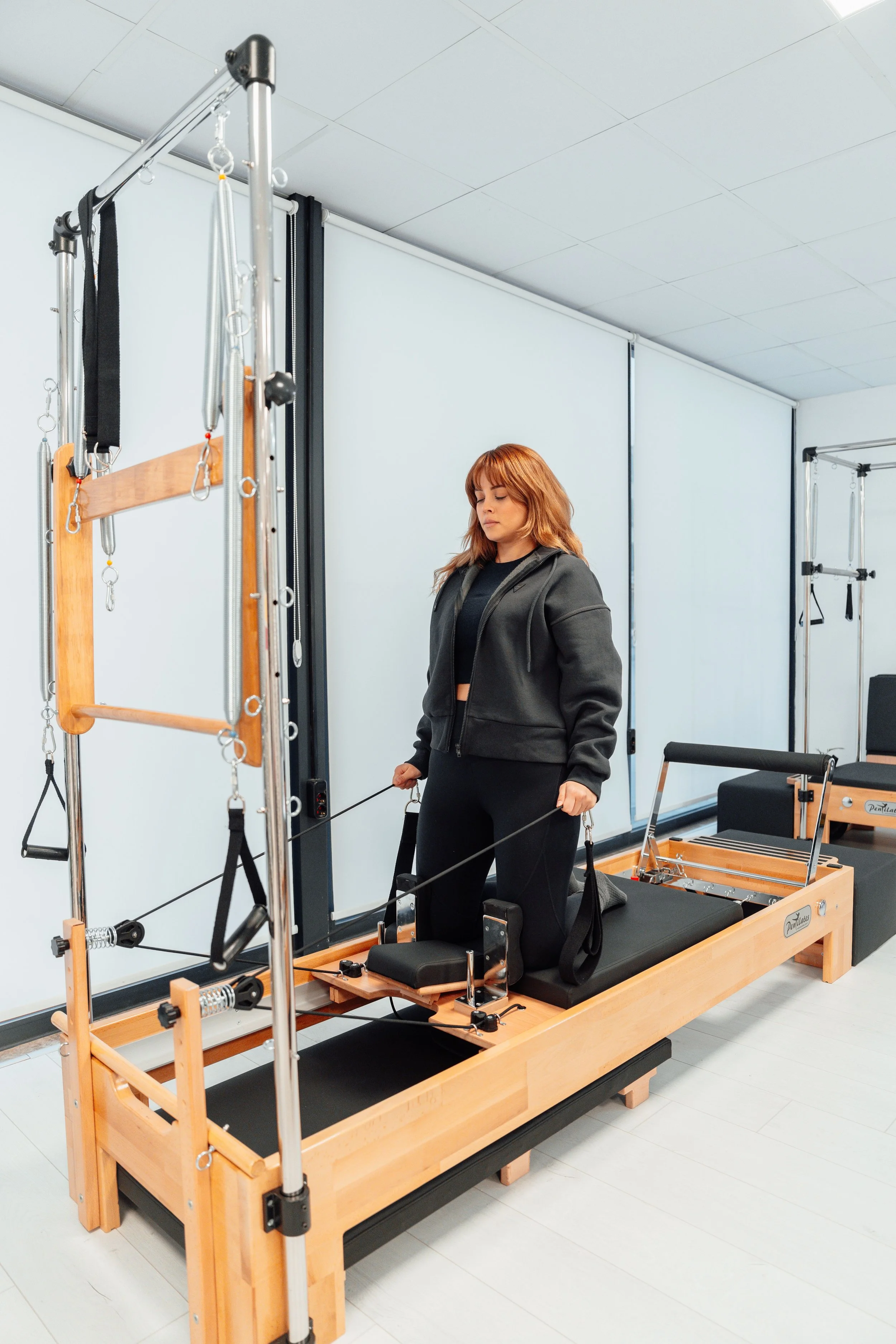 A woman standing on a Pilates reformer machine with various exercise equipment around her in a fitness studio.
