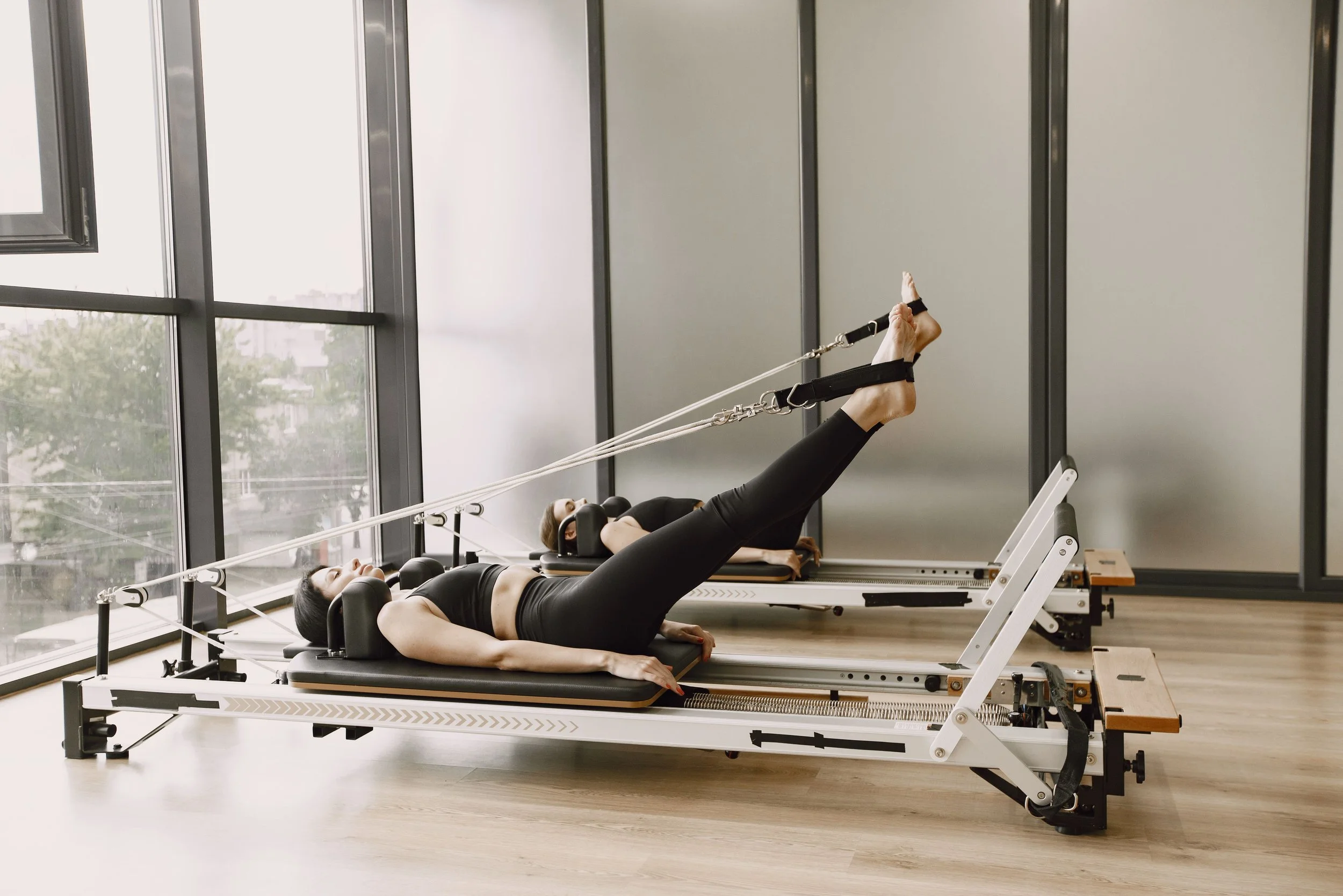 Two women lying on Pilates reformer machines, engaging in Pilates exercises, with large windows and cityscape view in the background.