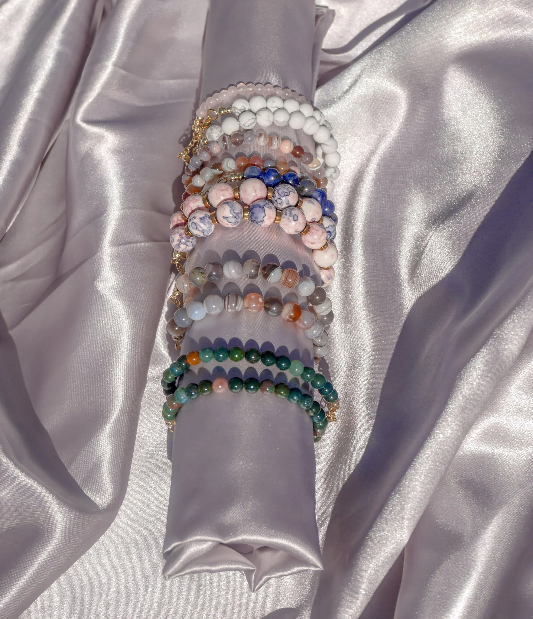 Multiple beaded bracelets in various colors and patterns are displayed on a gray satin fabric. The bracelets include white, pink, blue, green, and multicolored beads, some with gold accents.