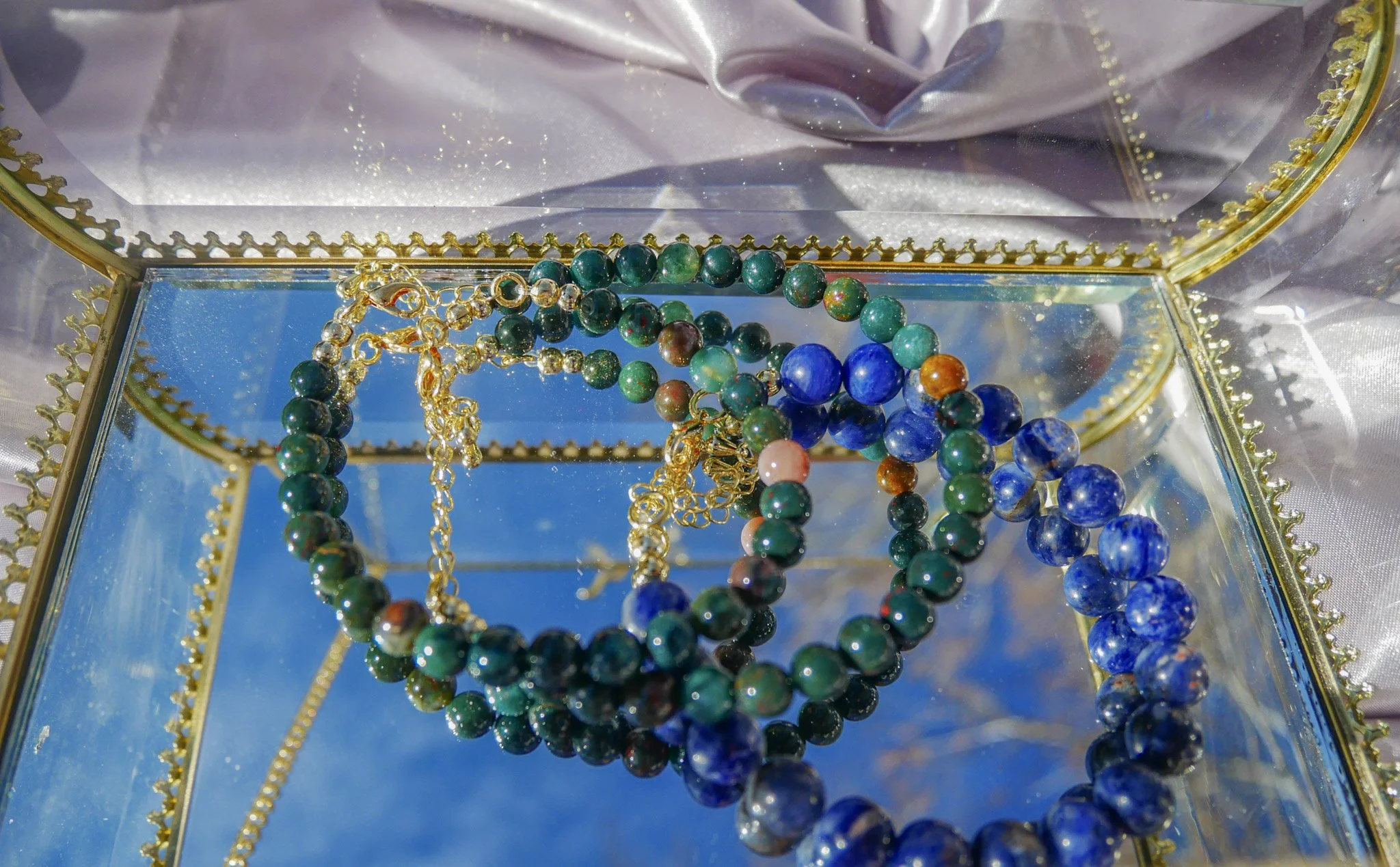 Colorful beaded jewelry, including necklaces and bracelets, displayed inside an ornate glass jewelry box with a mirrored lid, with a partly visible satin fabric background.