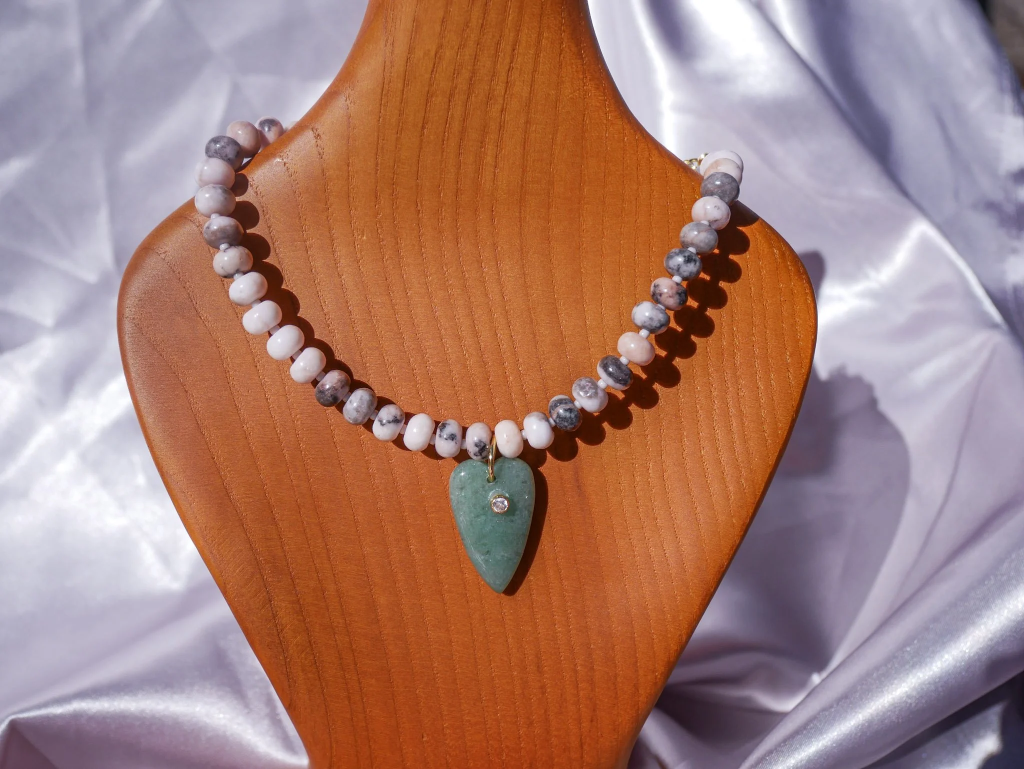 Beaded necklace with a green heart-shaped pendant on a wooden display bust, satin fabric background.
