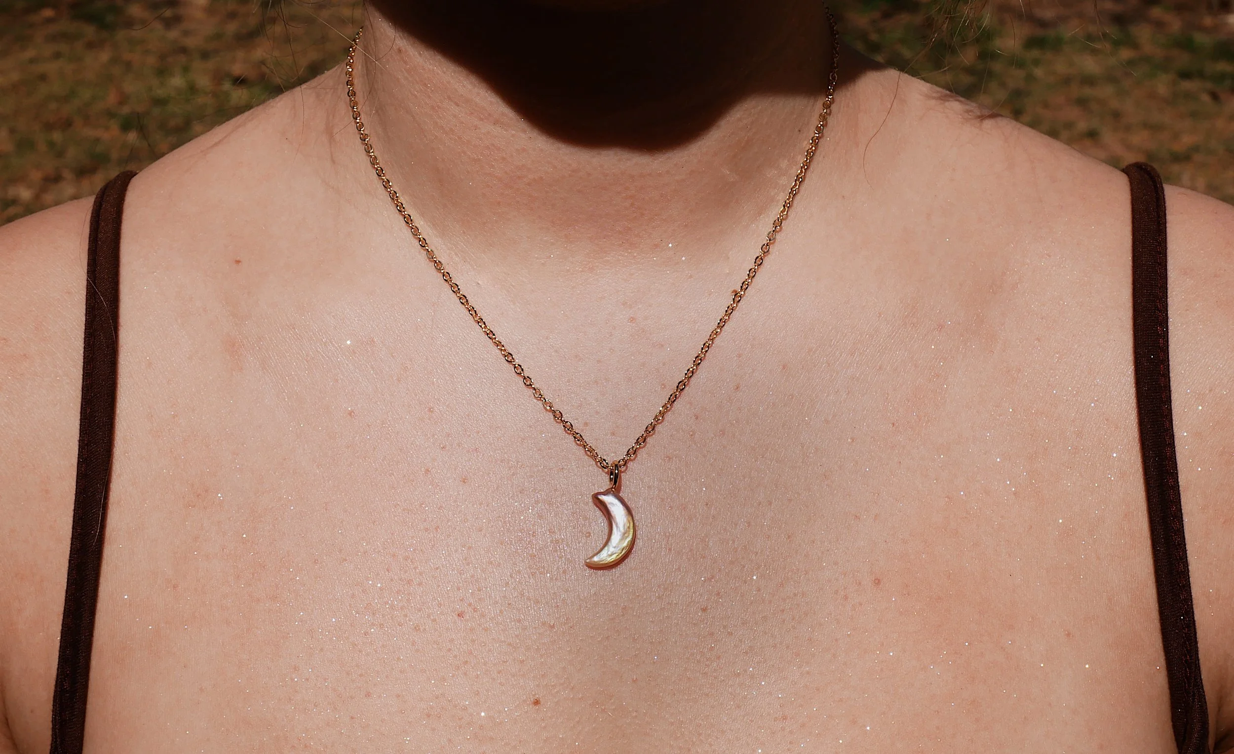 Close-up of a woman's upper chest wearing a gold chain necklace with a moon-shaped pearl pendant, in an outdoor setting.