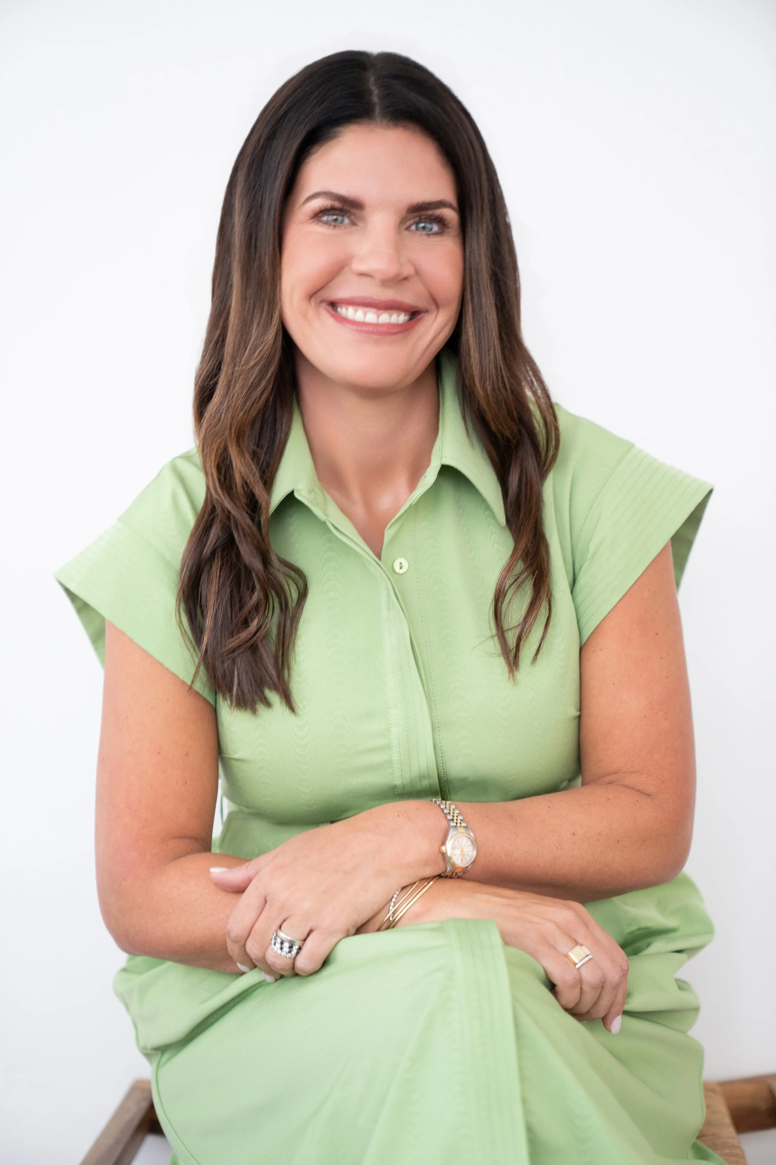 A woman with long brown hair, smiling, wearing a light green dress, sitting with her arms crossed in front of her, against a white background.
