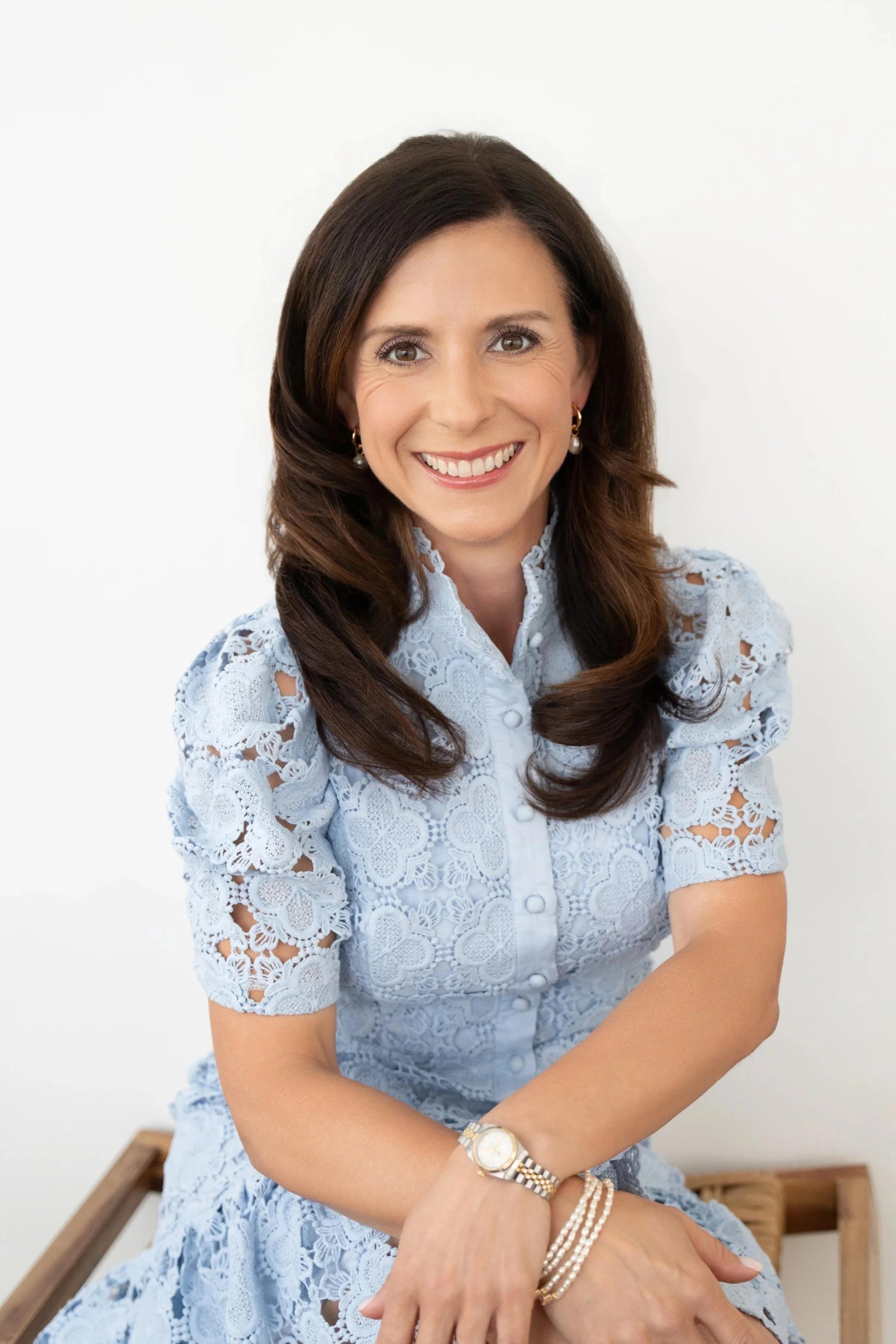 Smiling woman with brown hair wearing a light blue lace dress and pearl jewelry, sitting against a plain white background.