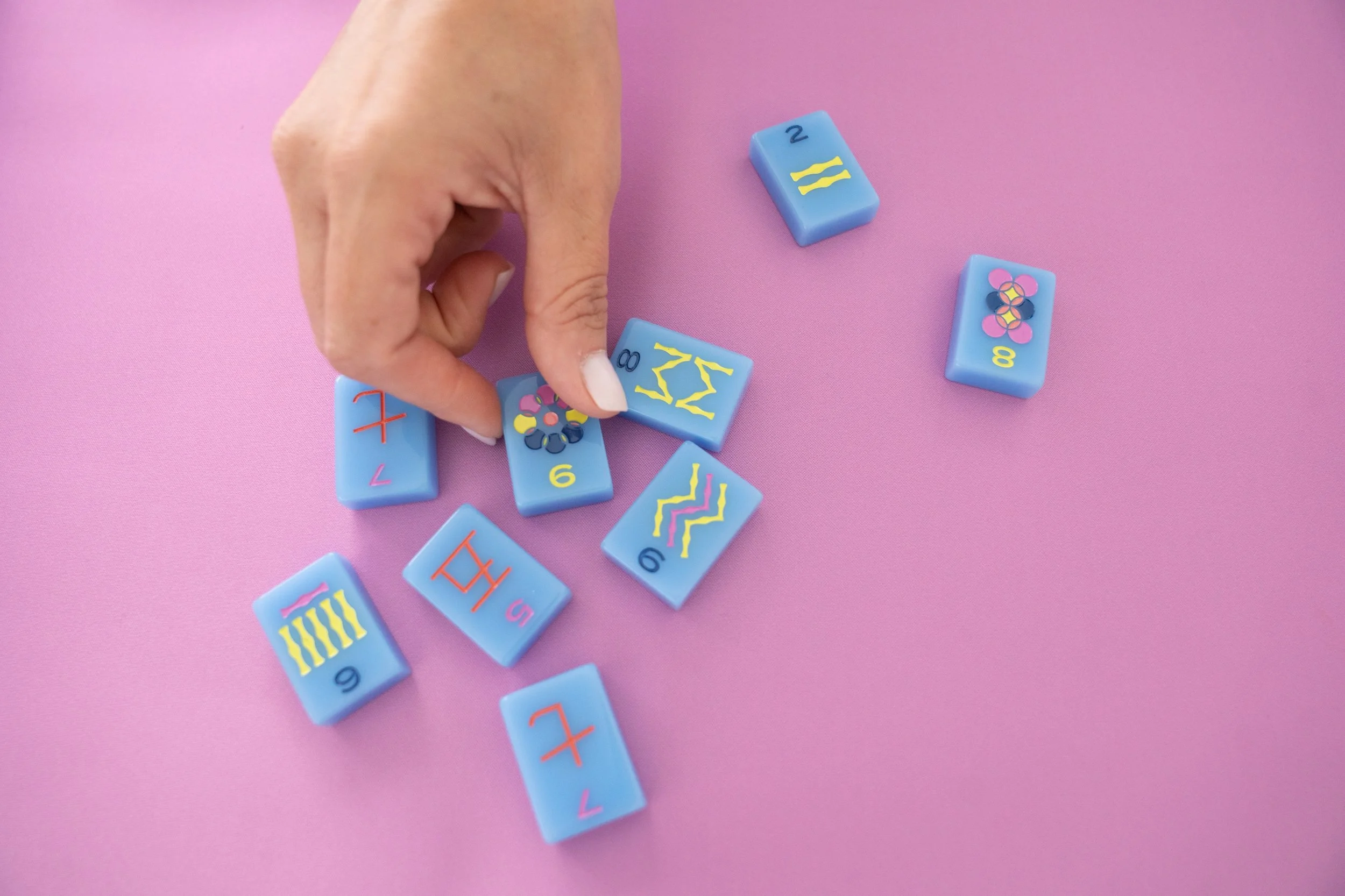 A hand arranging colorful domino-like tiles on a pink surface. The tiles are blue with various colored numbers and abstract patterns.