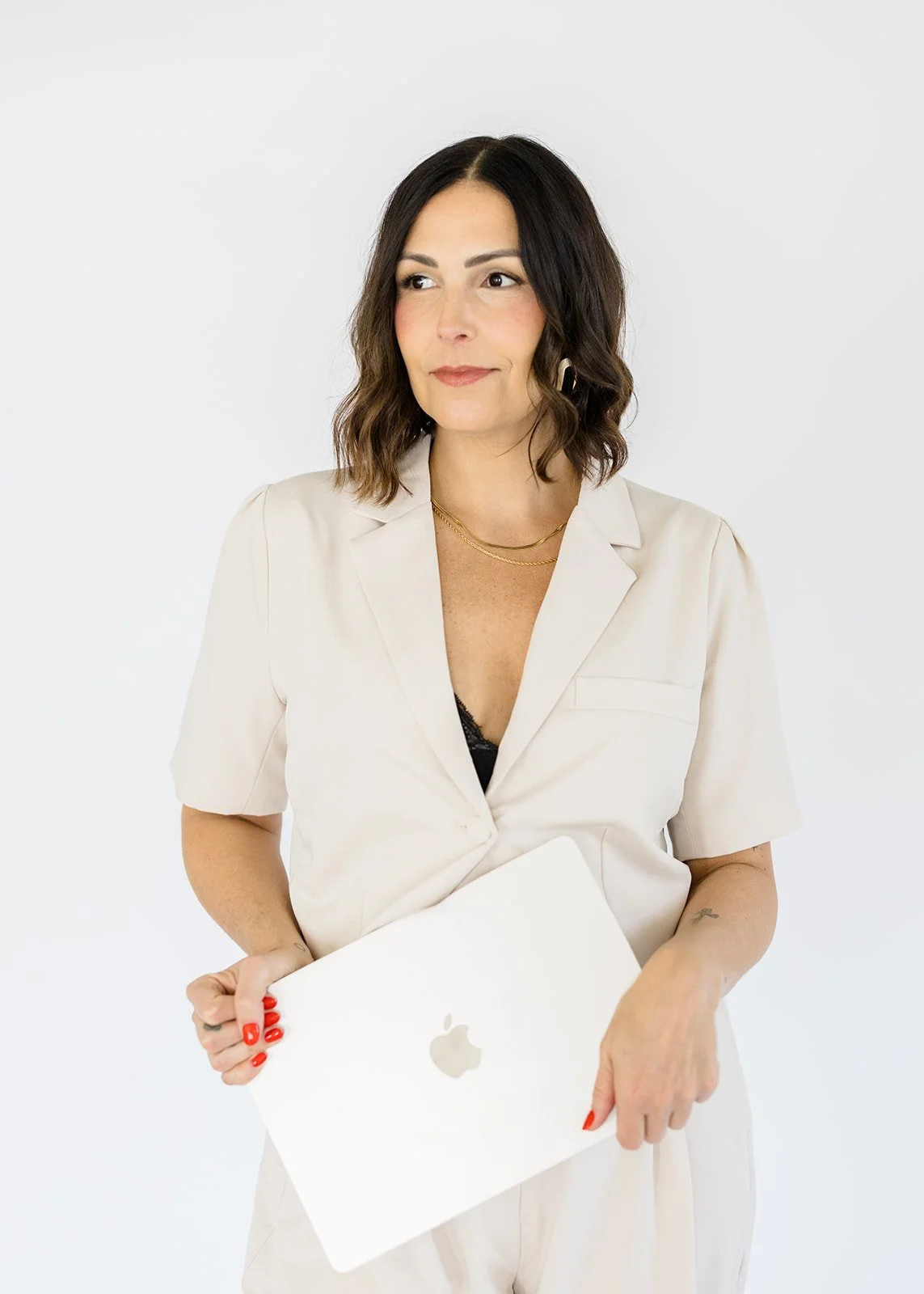 A woman with shoulder-length dark hair, wearing a light beige blazer and holding a closed white Apple MacBook, looking to her right against a plain white background.