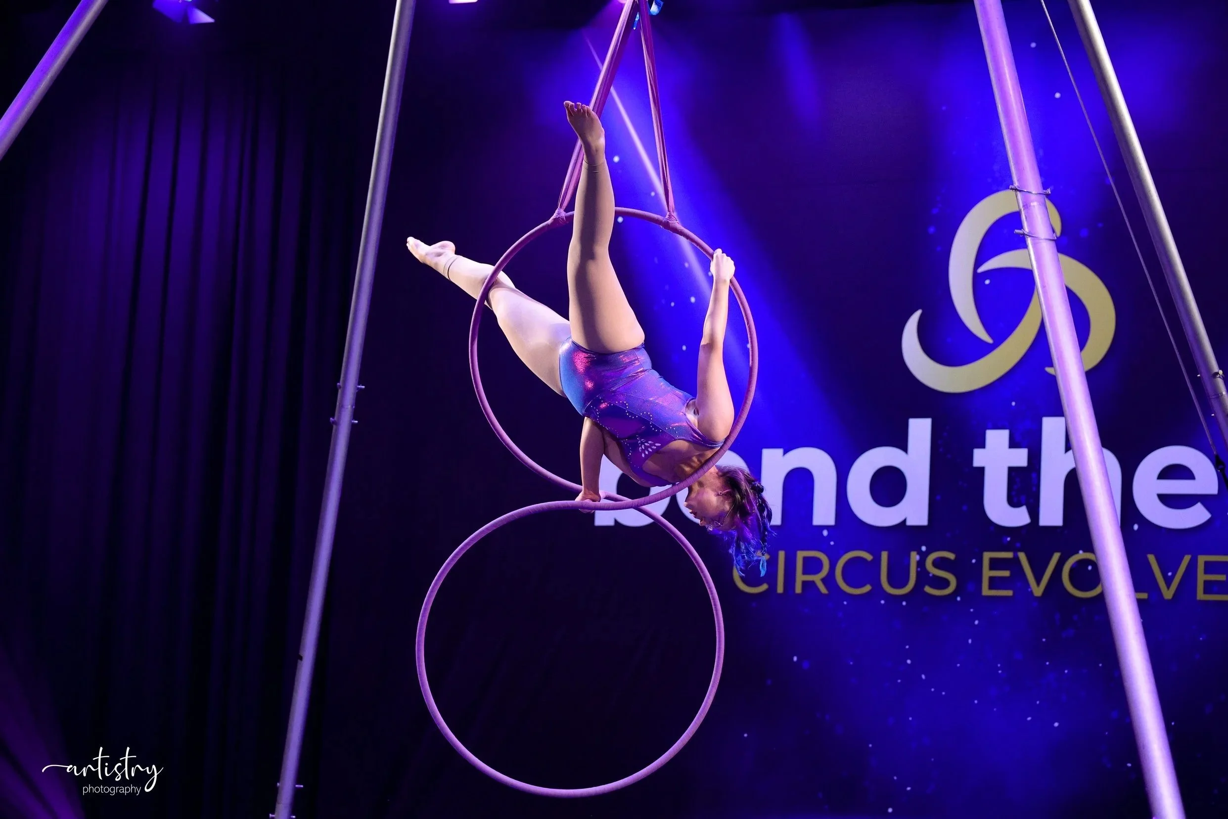 A female aerialist performing on pink gymnastic rings suspended from a metal frame, wearing a shiny purple leotard with star patterns, in a dark setting with purple lighting and a backdrop with the words 'Circus Evo'