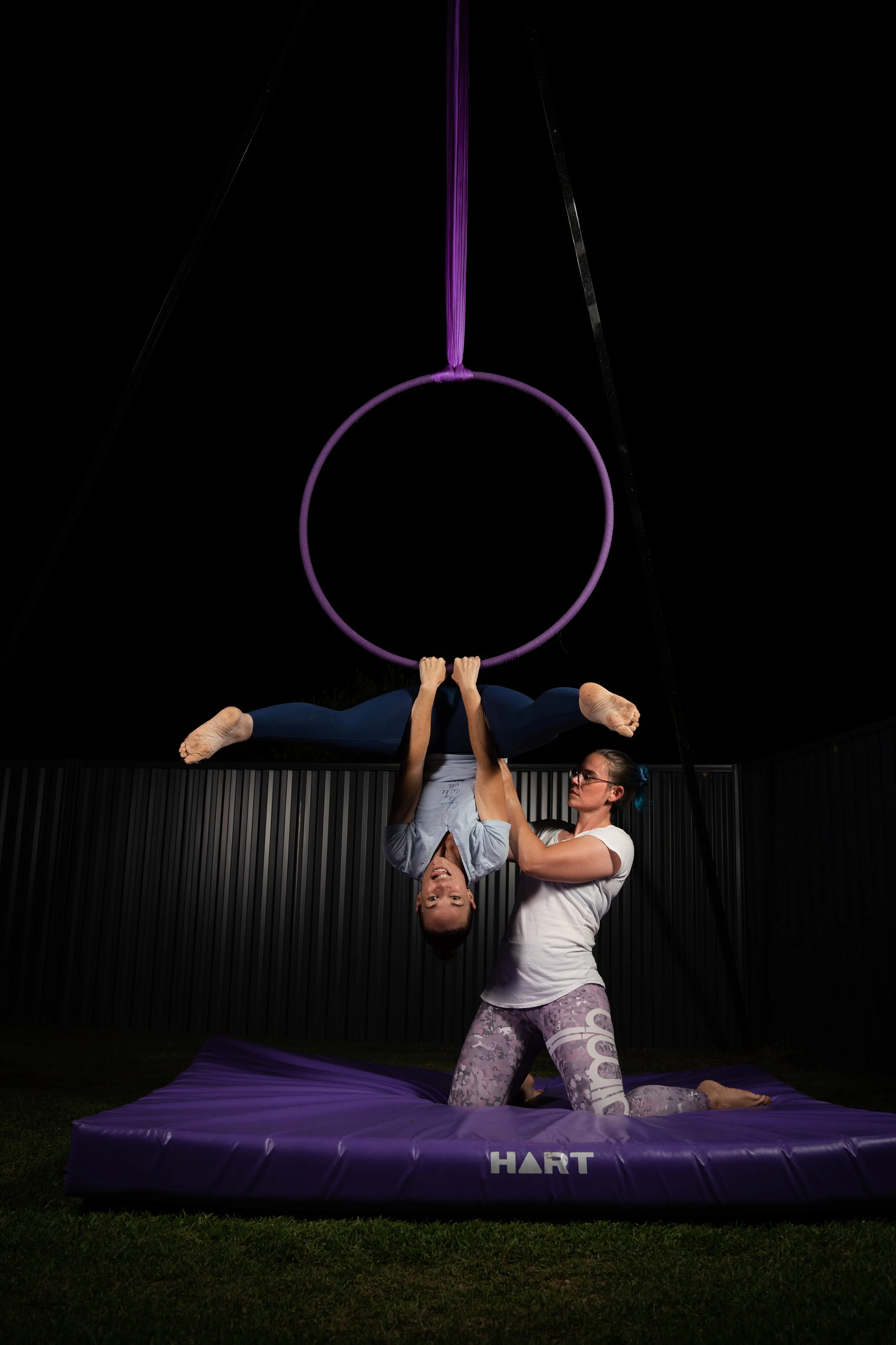 A woman is performing an aerial acrobatics move on a purple trapeze suspended from above, while another woman supports her from below on a purple cushioned mat outdoors at night. The background features a dark sky and a corrugated metal fence.