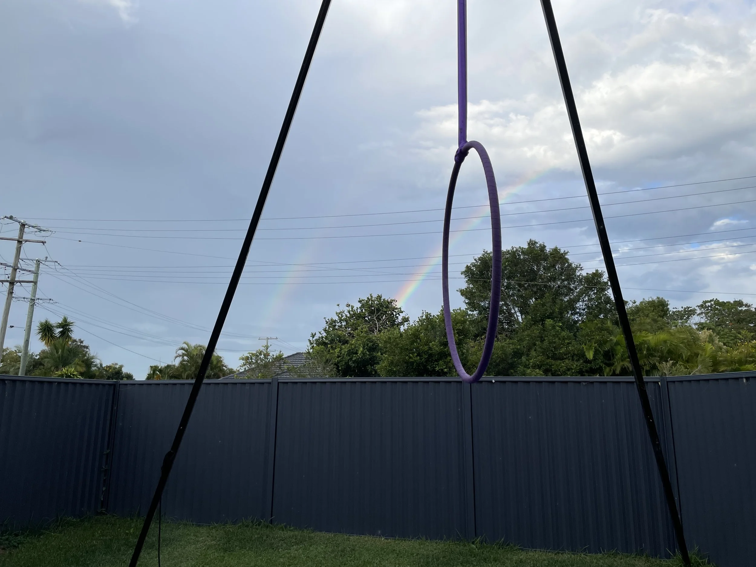 A purple trampoline hoop in the foreground of a cloudy sky, rainbow, power lines, trees, and a fence in the background.
