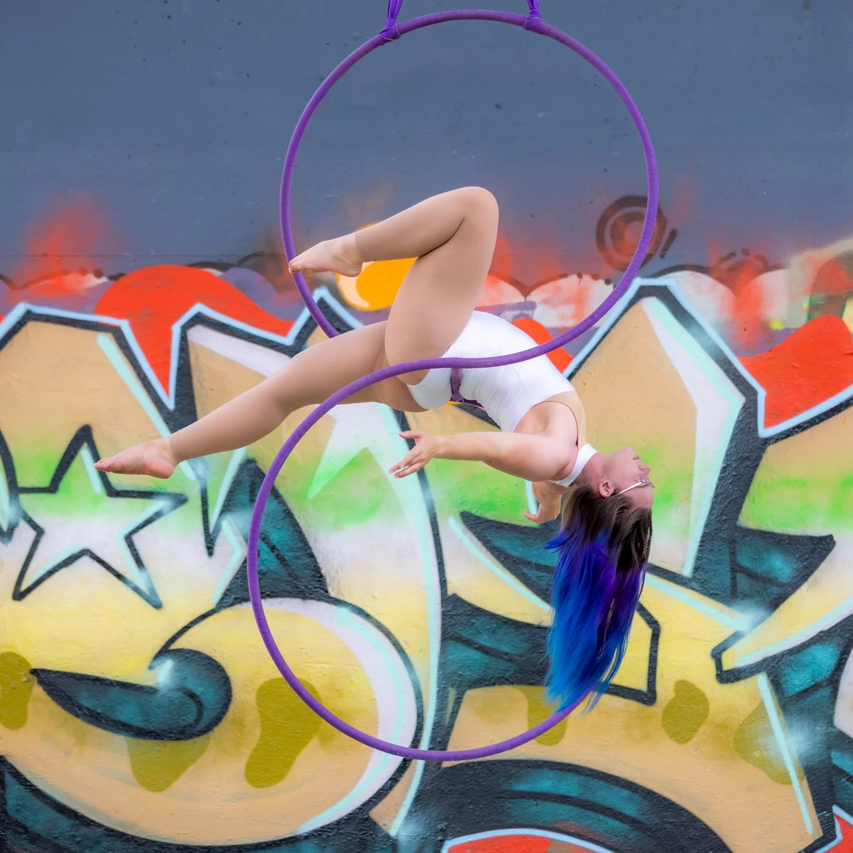 A woman with long blue and purple hair performing aerial acrobatics with a purple hoop in front of a colorful graffiti wall.