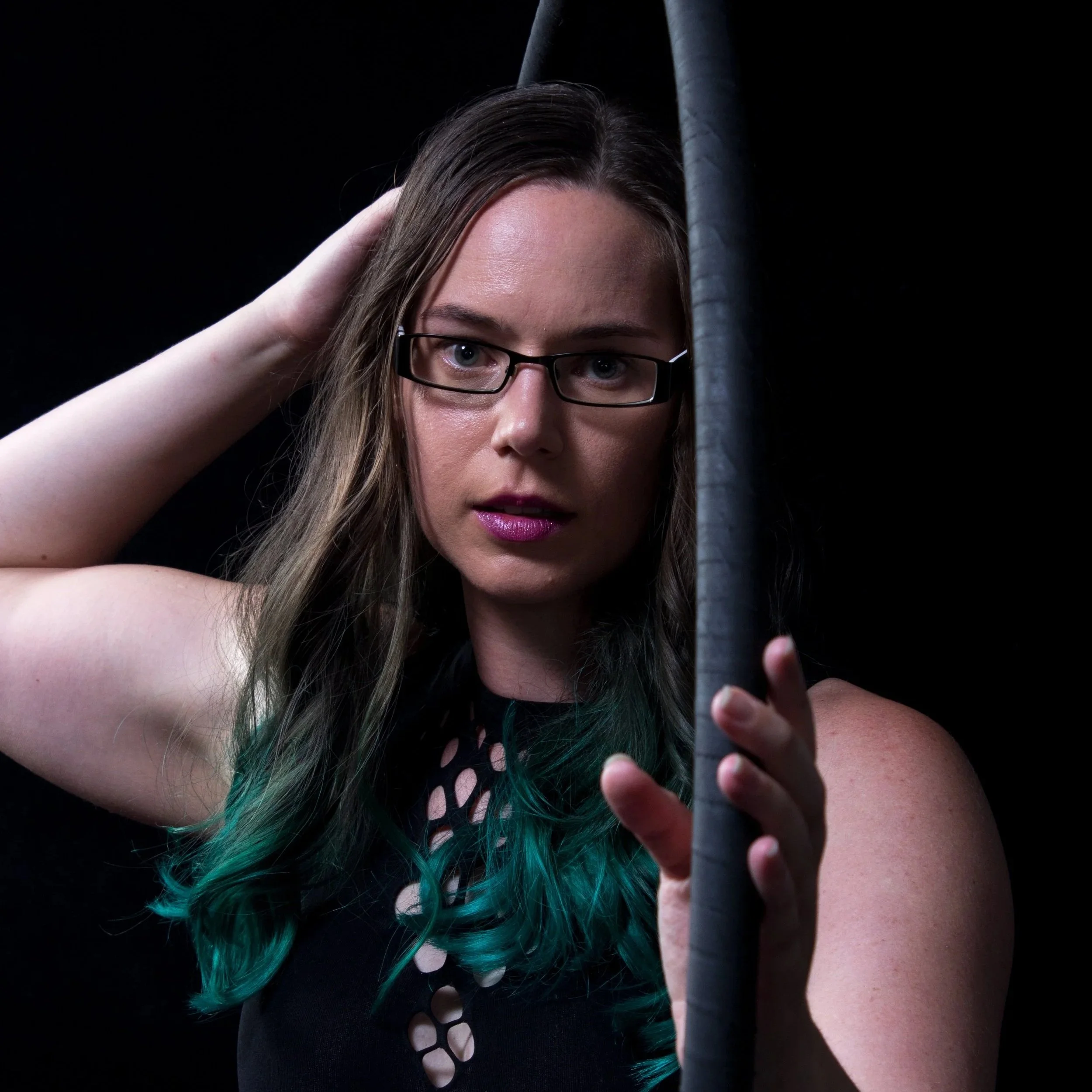 Close-up of a woman with long brown hair with green tips, wearing black glasses, reaching up with her right hand, holding a gray pole, against a dark background.