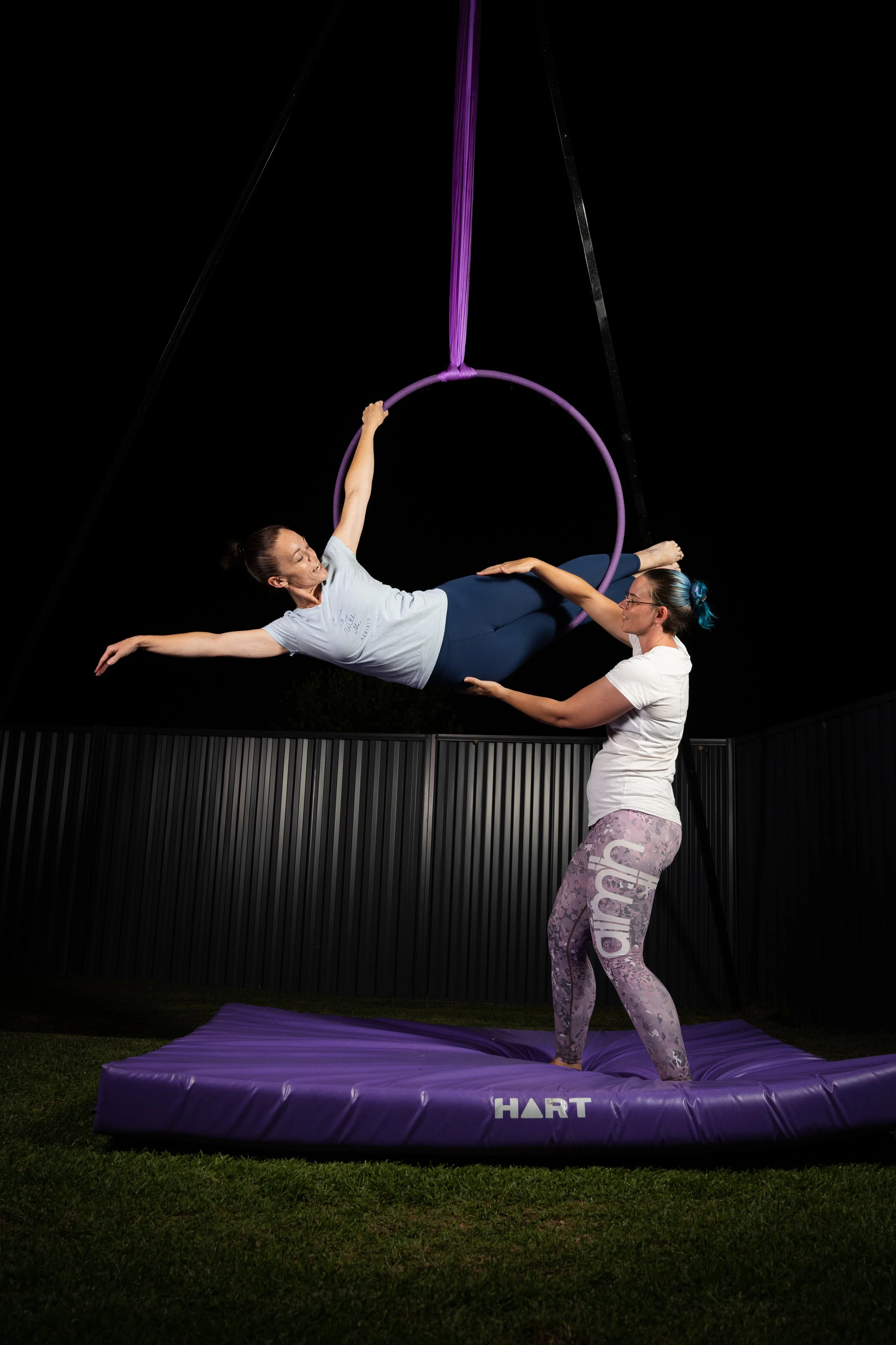 Two women practicing aerial acrobatics at night on a purple aerial hoop. One woman is holding the hoop and lying horizontally, while the other supports her and stands on a purple mat. They are outdoors in a fenced area with a dark sky.