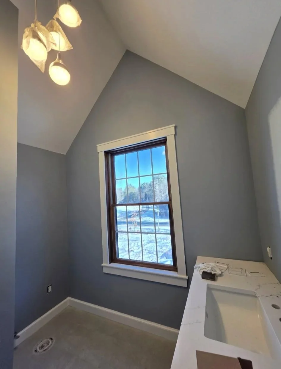 An unfinished bathroom with light blue walls, a large window with white trim showing a snowy outdoor scene, with a ceiling light fixture and a white countertop with a sink installation in progress.