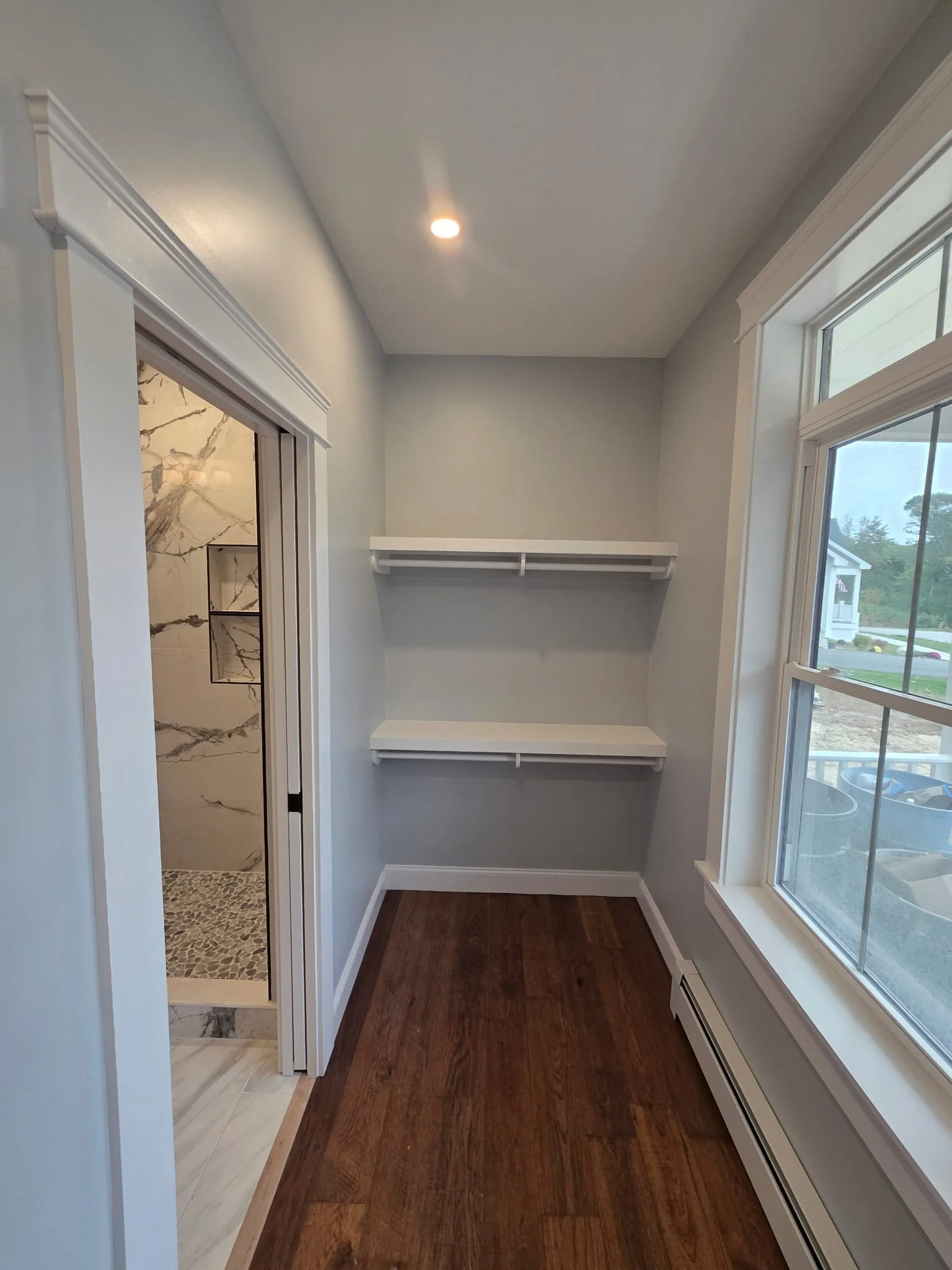 A small room with hardwood flooring, two white shelves on the back wall, a large window on the right, and a door leading to a bathroom with marble tiles and a pebble shower floor.