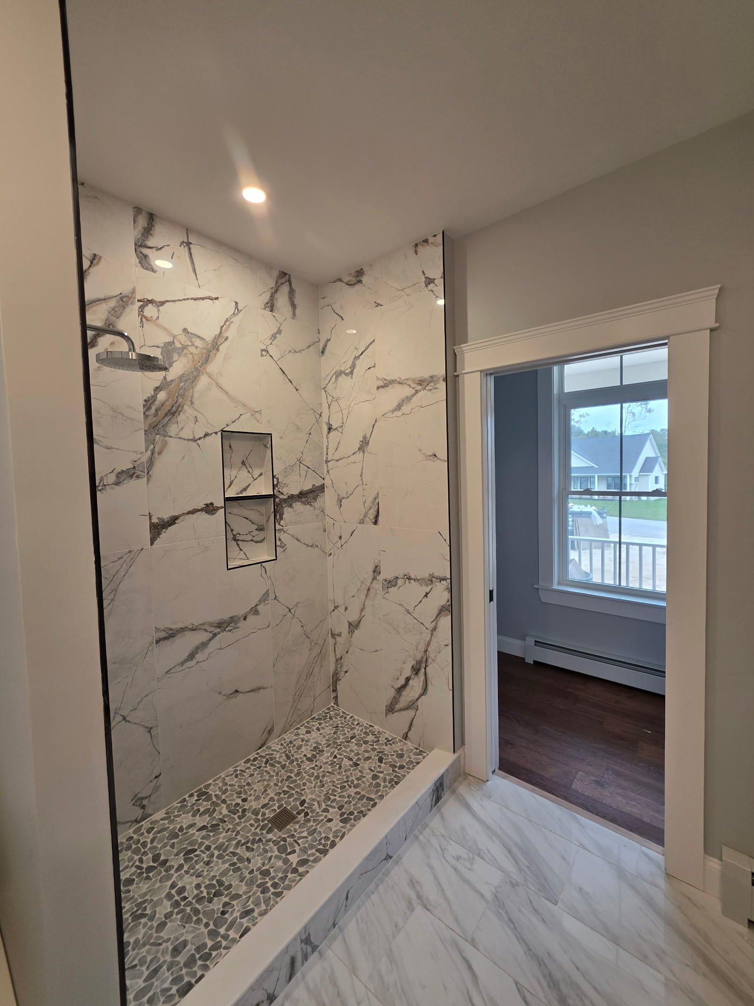 Modern walk-in shower with marble tiles, pebble floor, built-in niche, and a window with a view to the outside.
