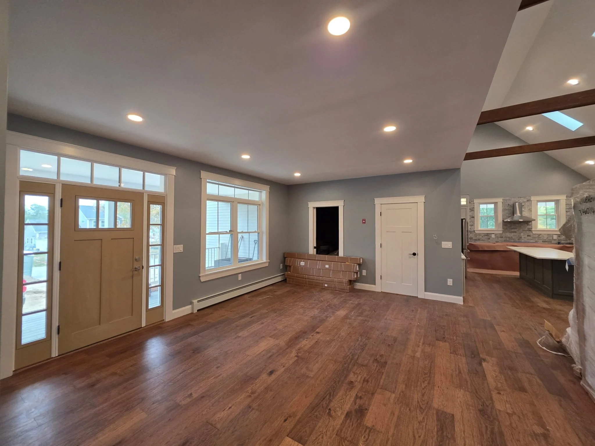 Interior of a modern living area with hardwood floors, large windows, recessed lighting, and an open kitchen in the background.