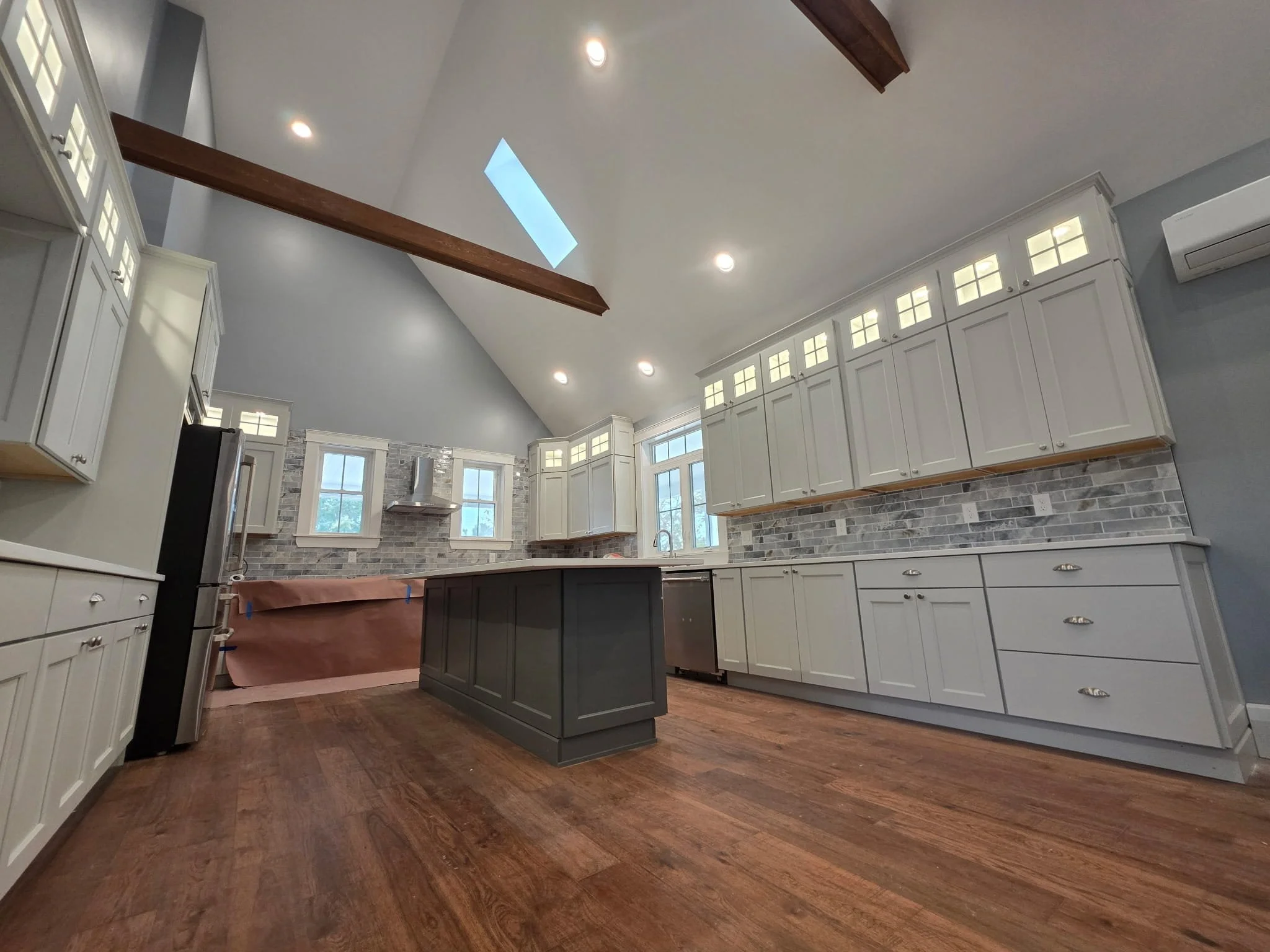 Kitchen under renovation with white cabinets, gray island, brick backsplash, hardwood floor, and skylight.