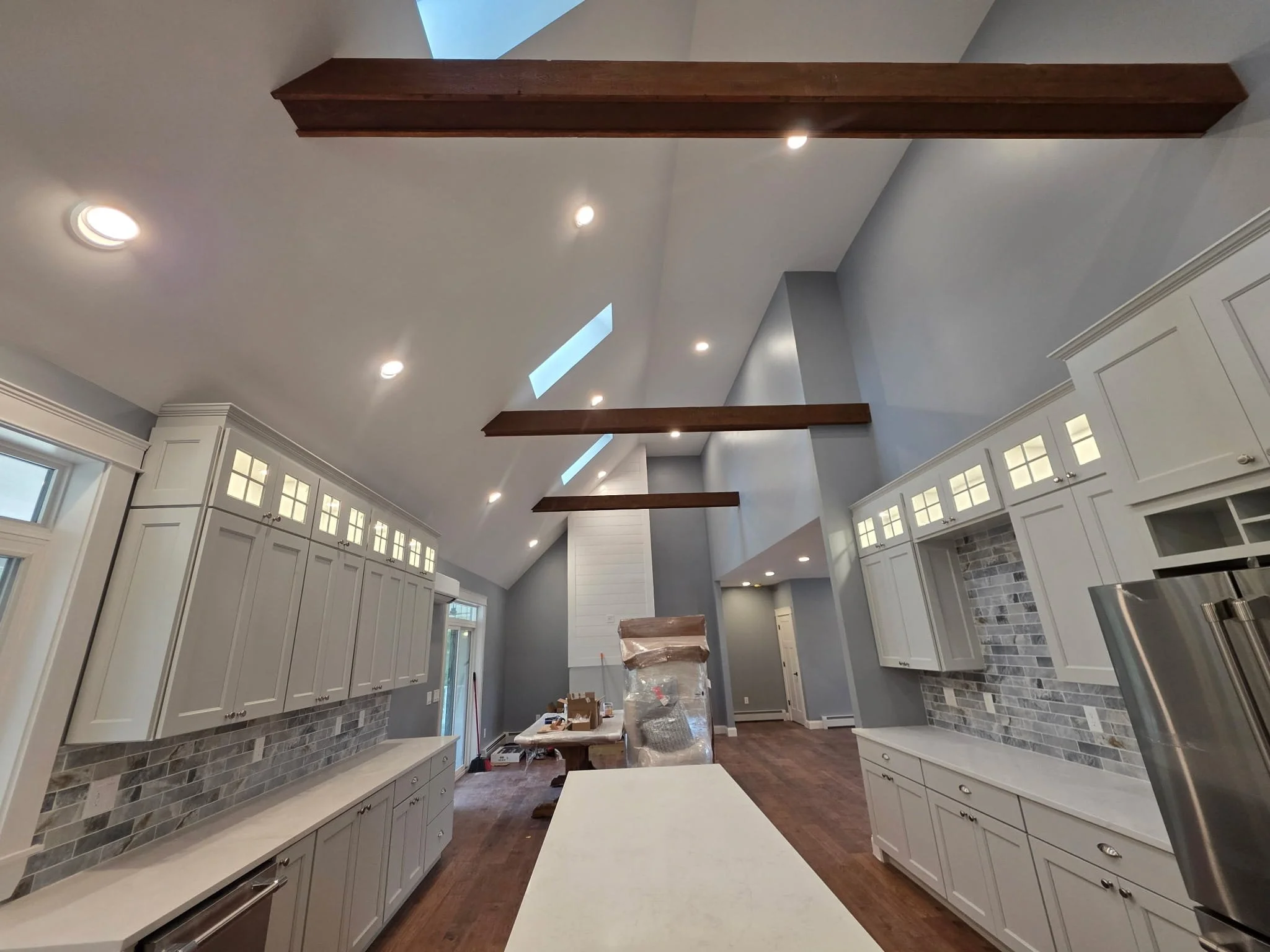 Modern kitchen with white cabinets, gray backsplash, hardwood floors, ceiling skylights, and exposed wooden beams.