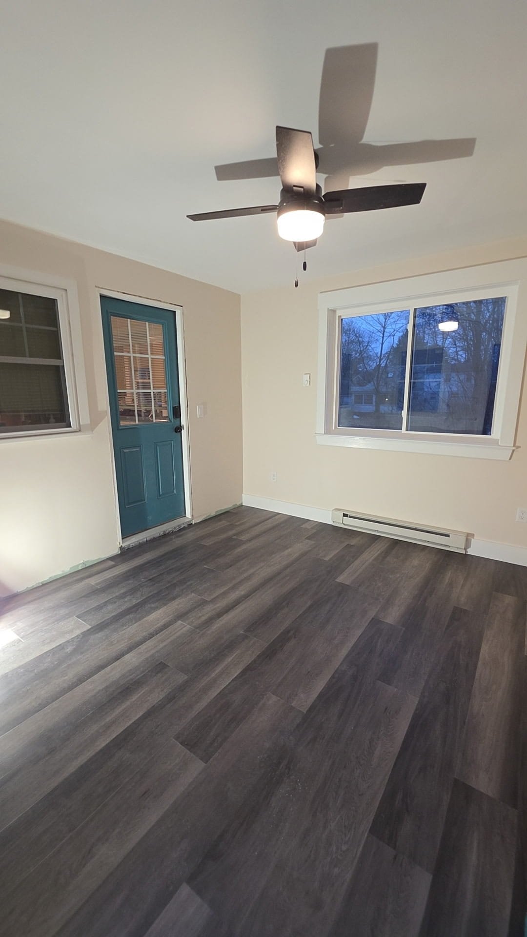 Empty room with dark wood flooring, white walls, a ceiling fan with a light, a large window, and a teal door.