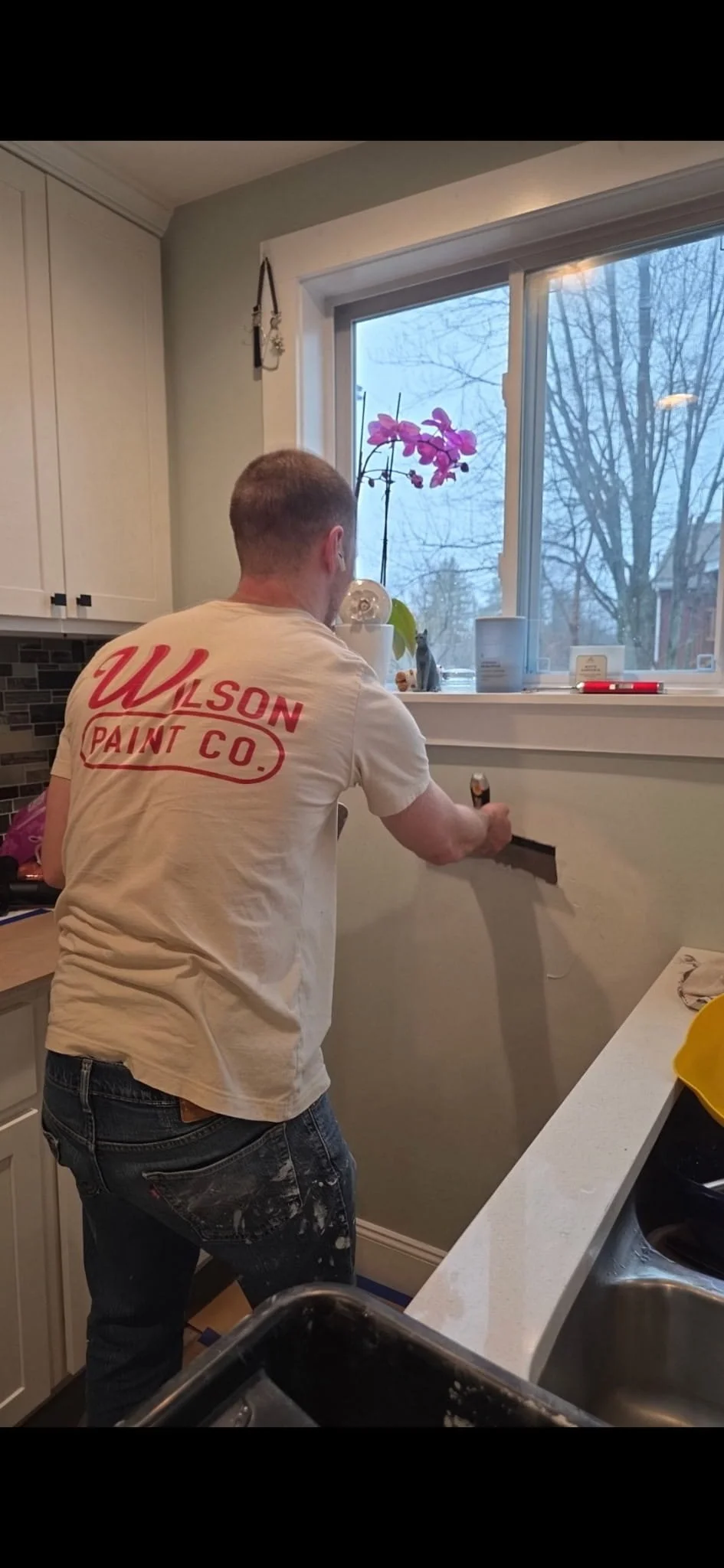 Man installing or repairing a wall outlet near a kitchen window, with tools and a purple orchid on the windowsill.
