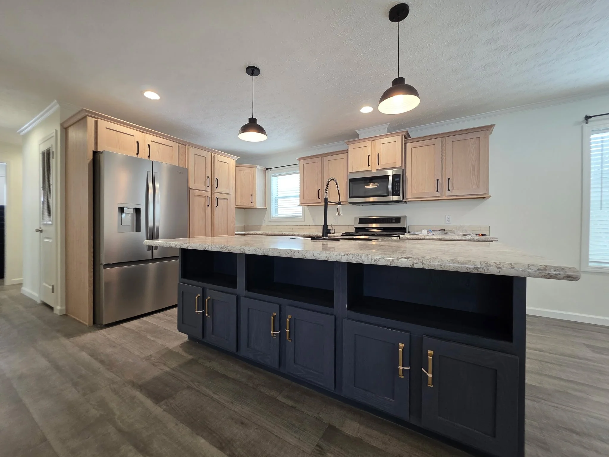 Modern kitchen with light wood upper cabinets, dark blue lower cabinets, stainless steel refrigerator, microwave, and stove, granite countertops, pendant lights, hardwood flooring, and windows bringing in natural light.