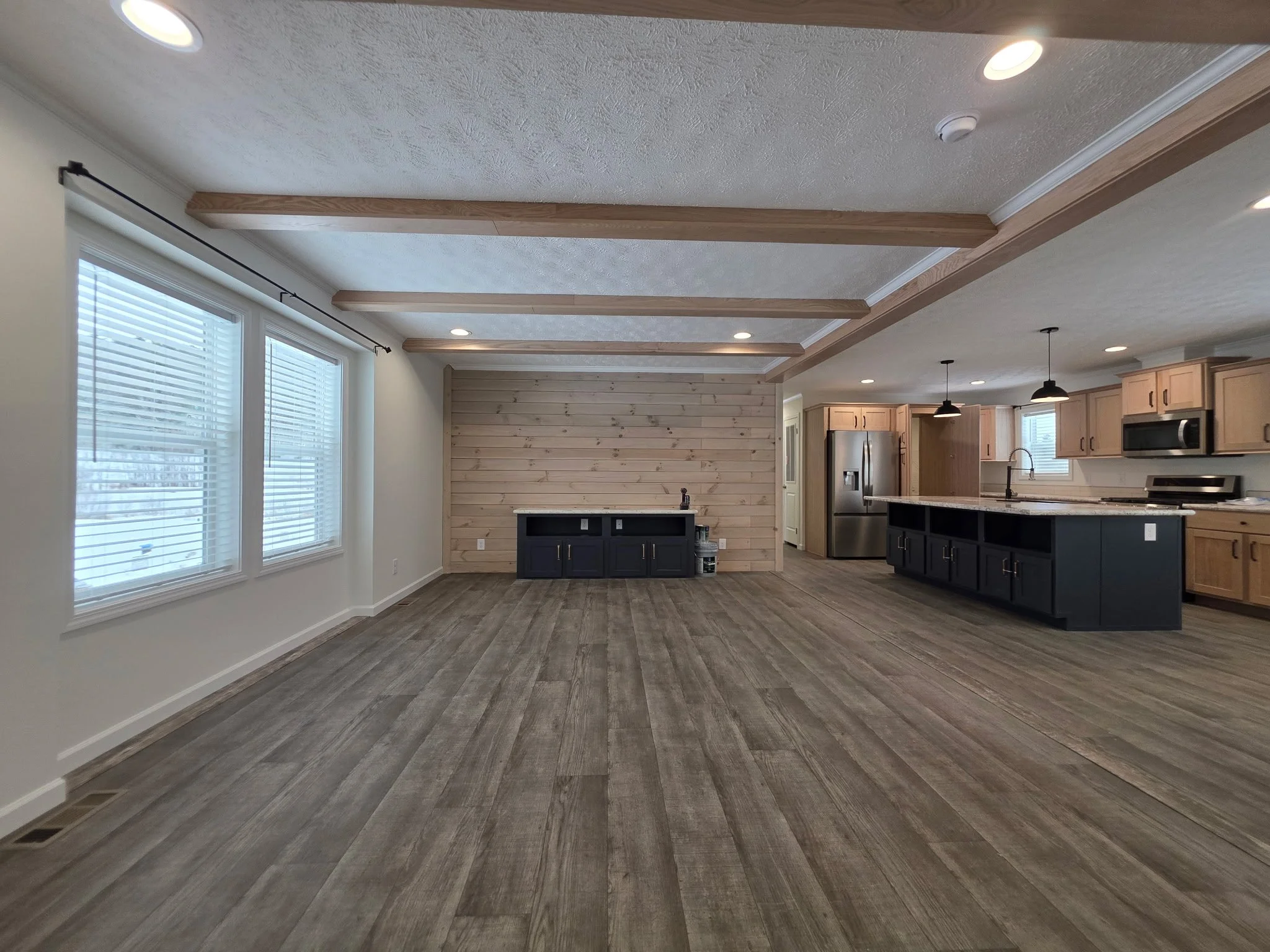 Empty living room and kitchen area with wood floors and ceiling beams, large windows with blinds, light-colored walls, a black lower kitchen cabinet island, and a wooden accent wall in the background.