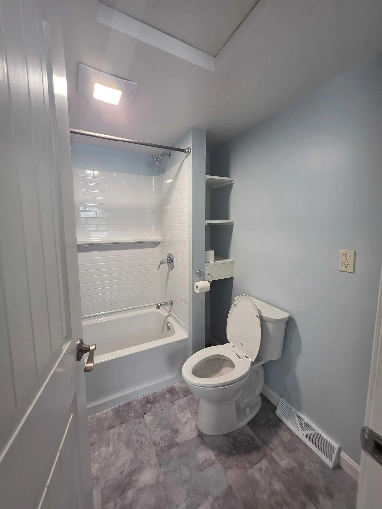 Small bathroom with bathtub and shower, toilet, light blue painted walls, gray tiled floor, built-in shelves, and a vent on the floor.