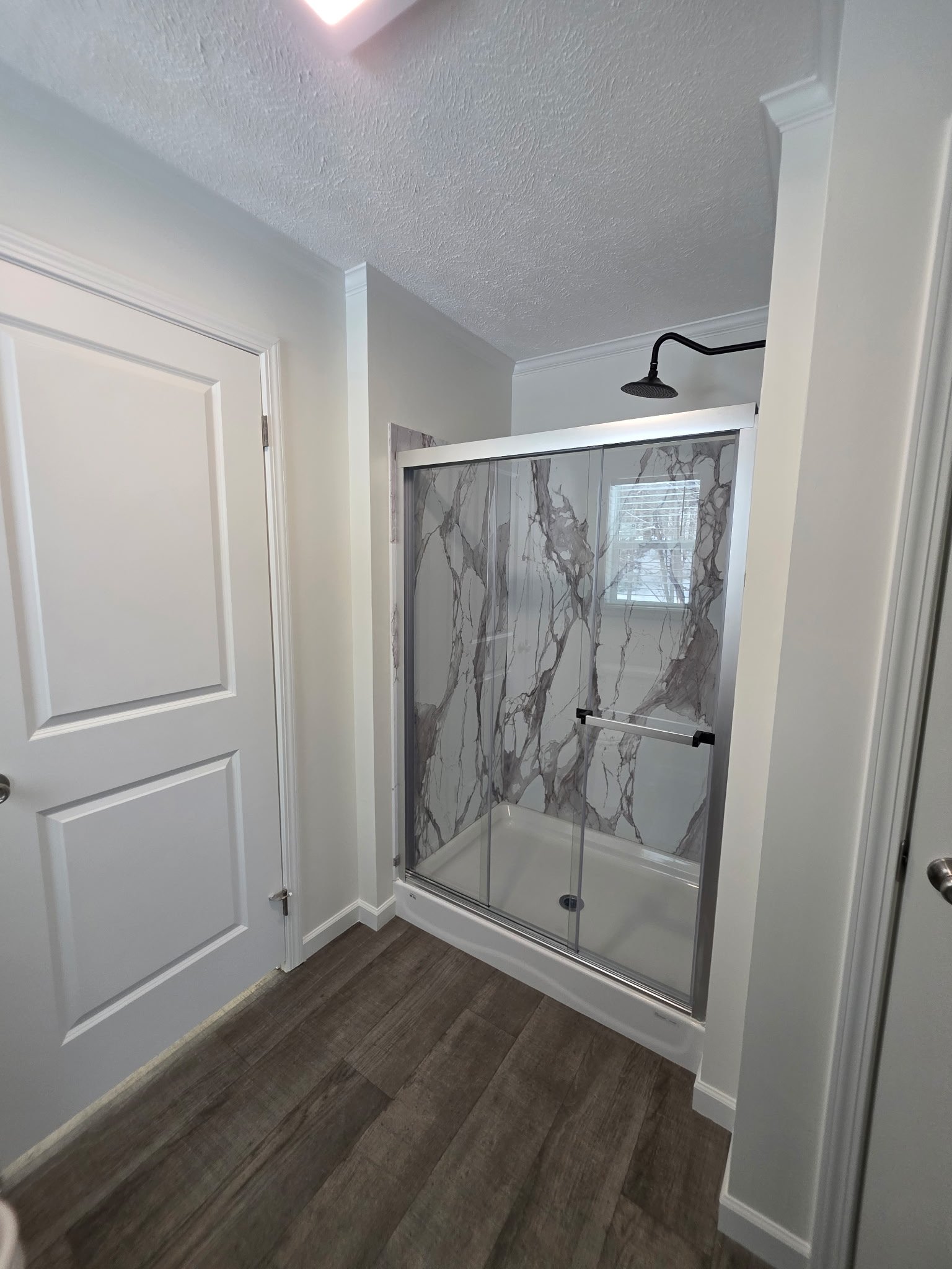Bathroom with a glass shower enclosure featuring a gray marble pattern, a black showerhead, white walls, wooden flooring, a white door, and a window with blinds.