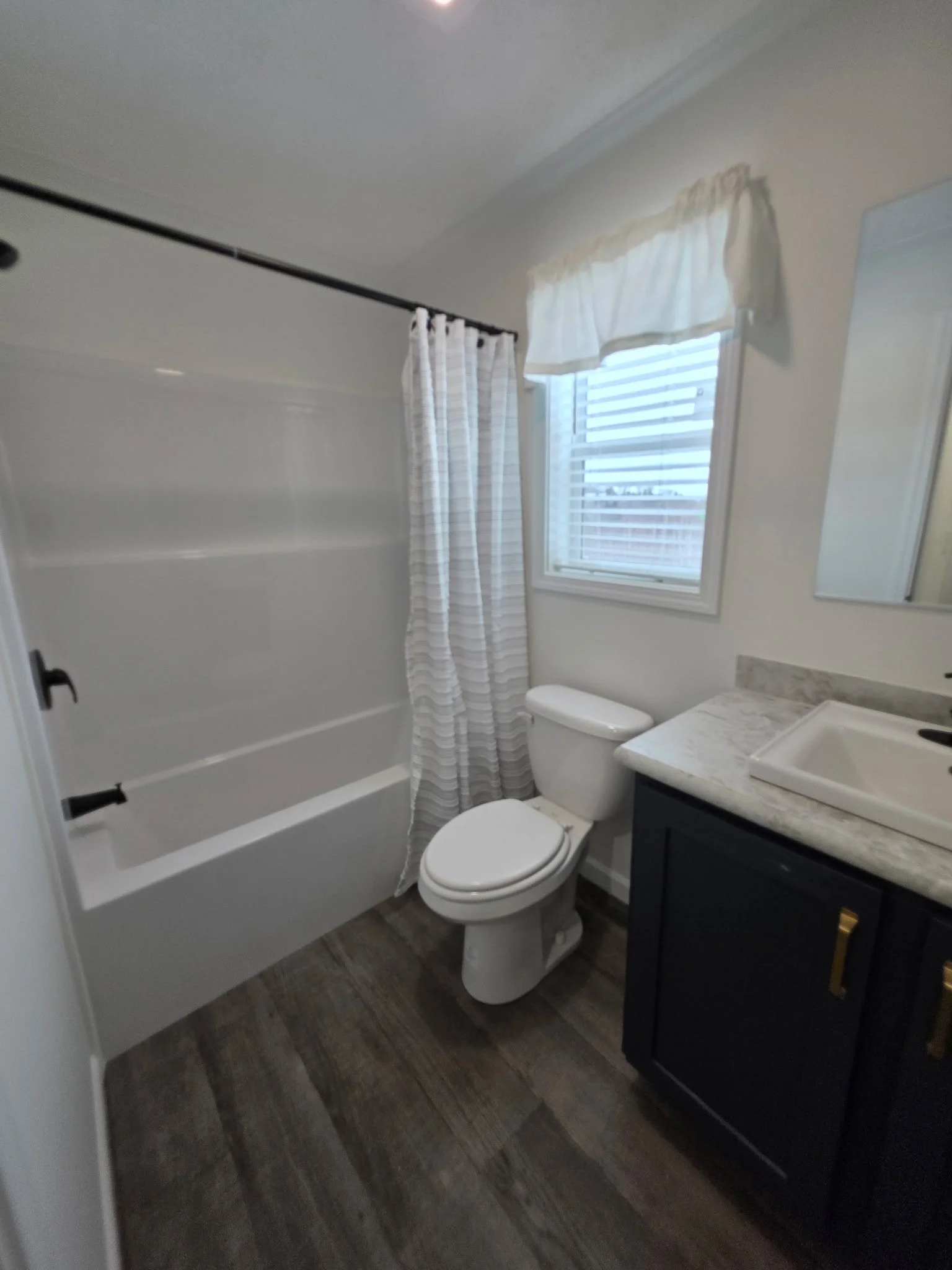 Bathroom with a white bathtub, toilet, dark vanity with a sink, window with blinds and a valance, wood-like flooring, and a mirror.