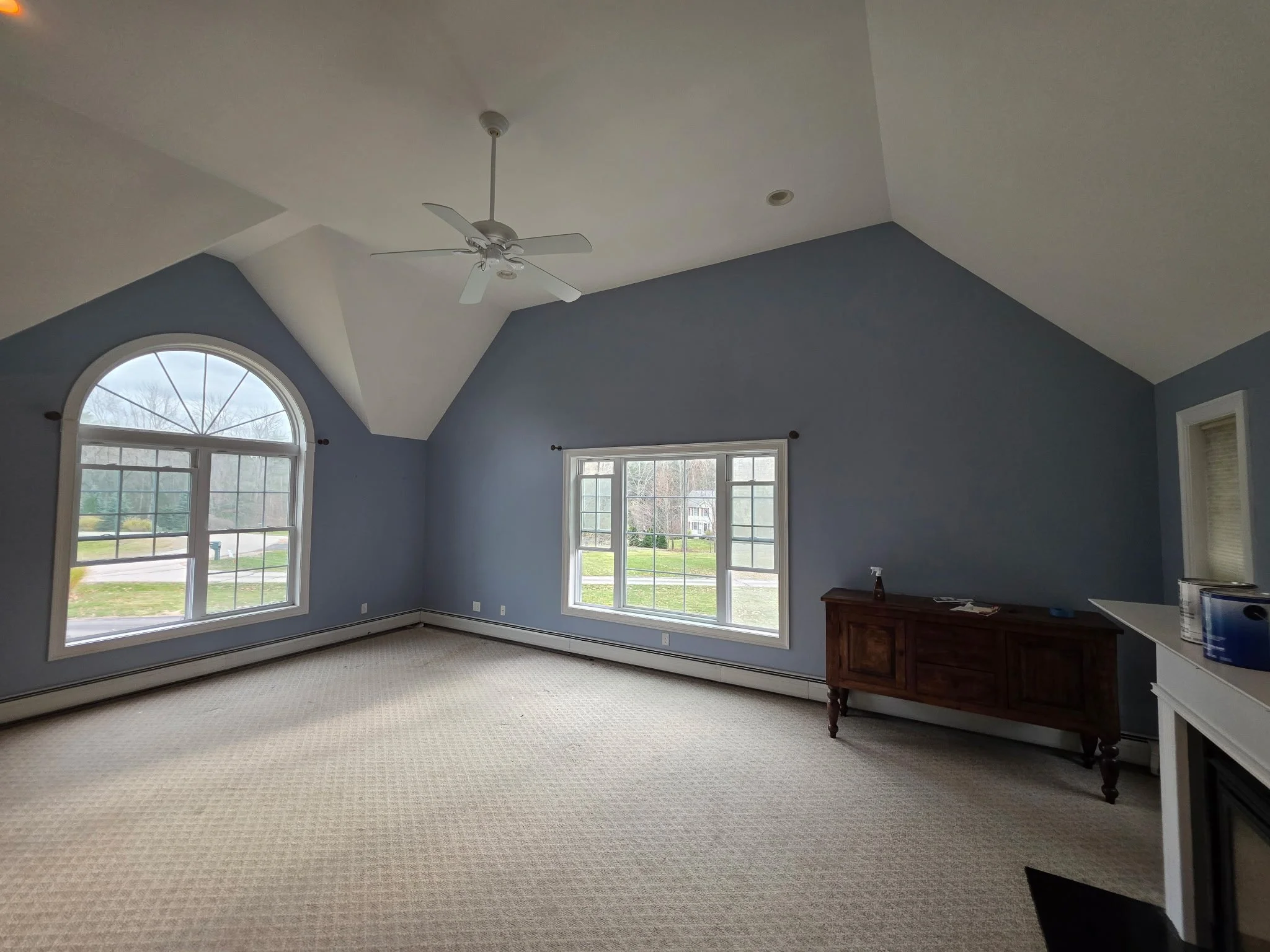 Empty living room with large windows, vaulted ceiling, ceiling fan, and beige carpet.