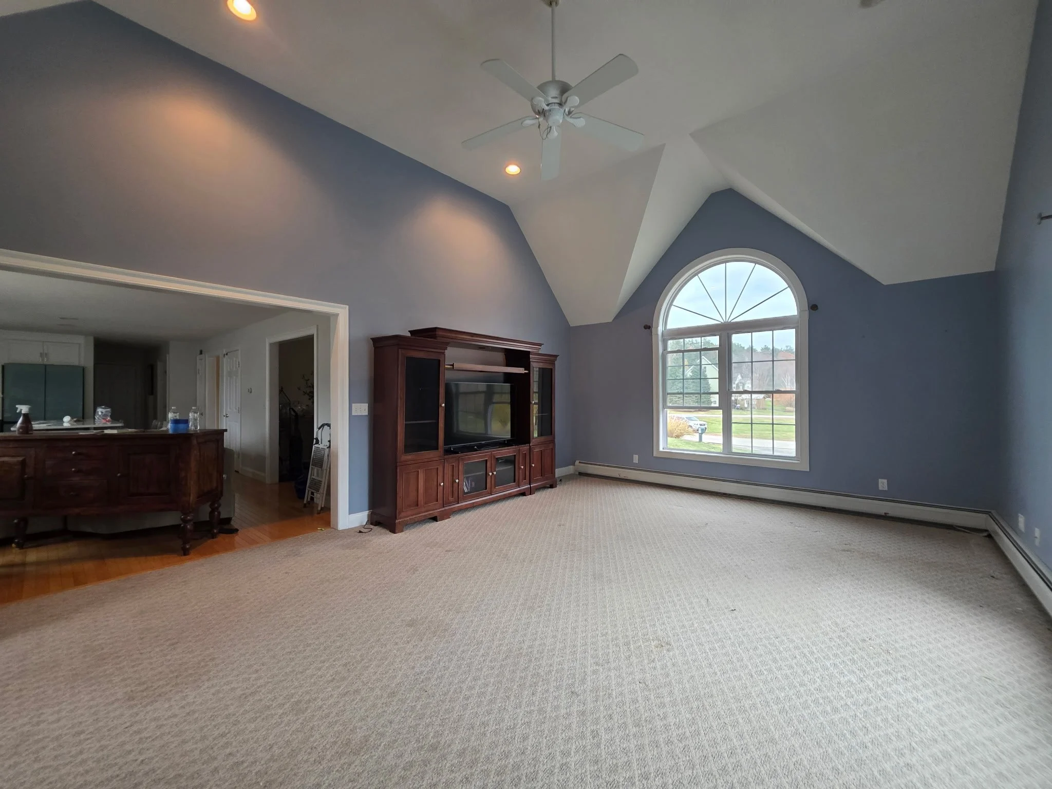 Empty living room with large arched window, light blue walls, beige carpet, and a dark wood entertainment center.