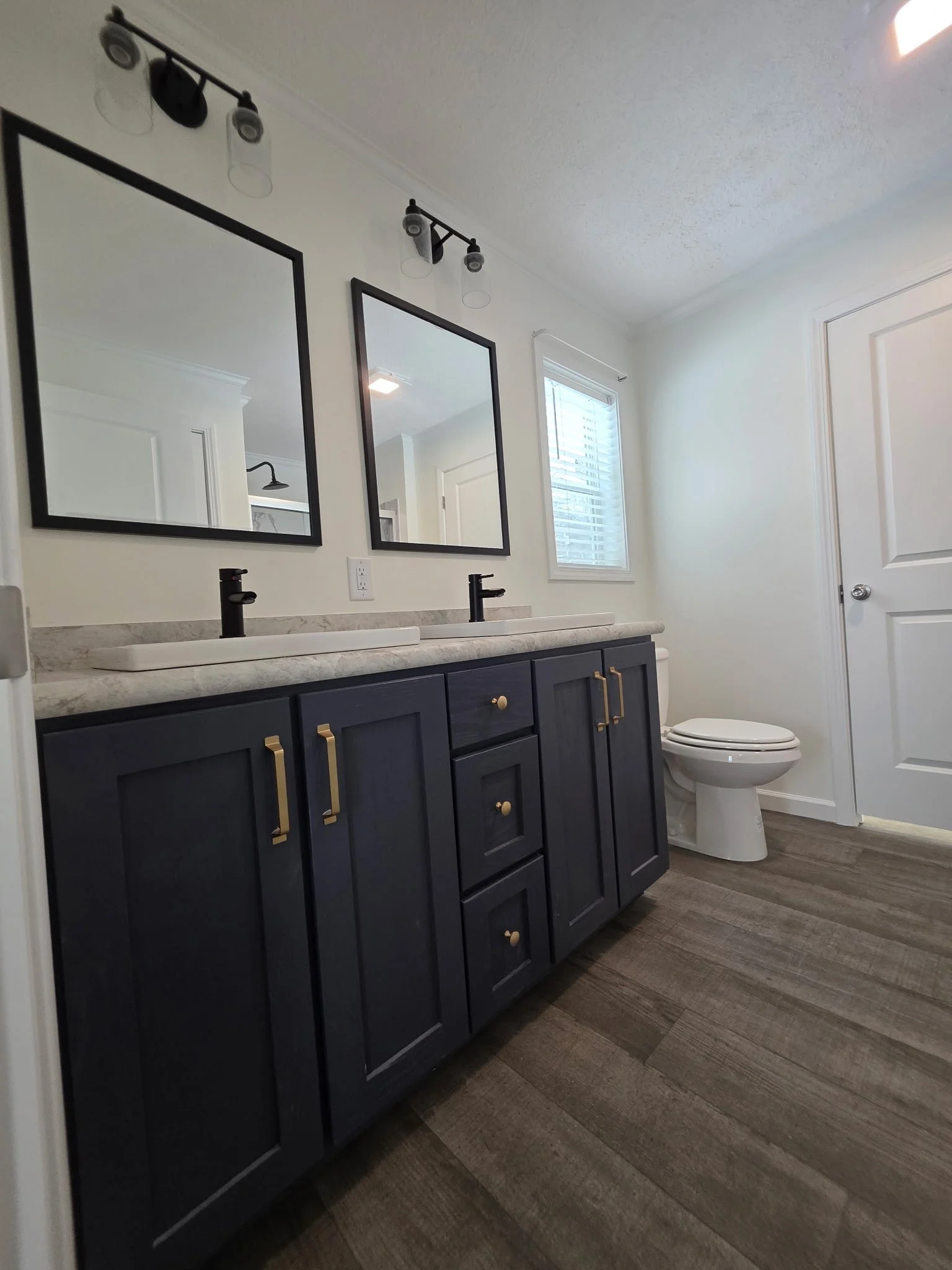 Bathroom with a double vanity with dark blue cabinets, black faucets, and two mirrors. There is a toilet and a window with blinds. The floor is wood-look tile.