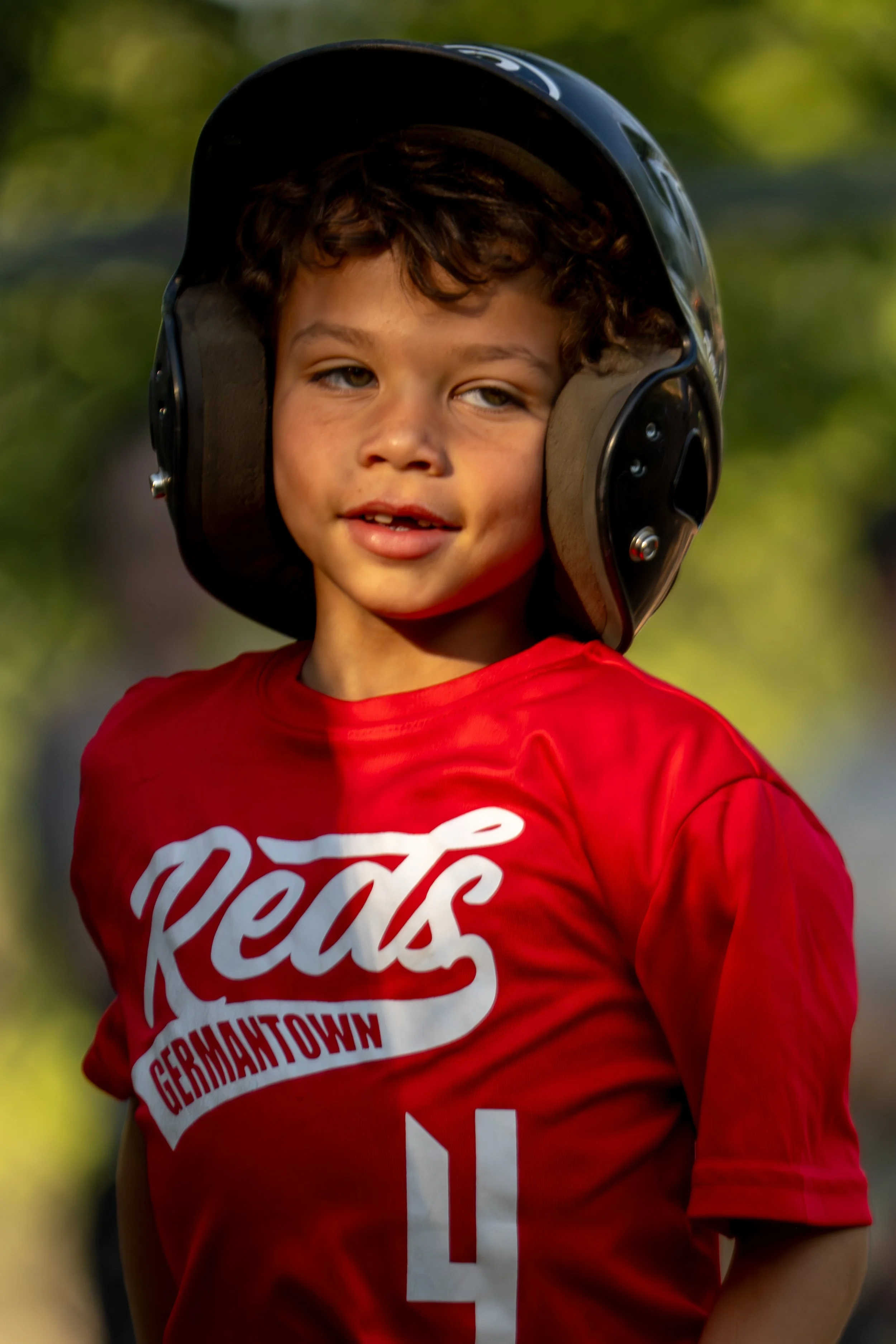 A young boy wearing a black helmet and a red t-shirt with white text and graphics, outdoors with a blurred green background.