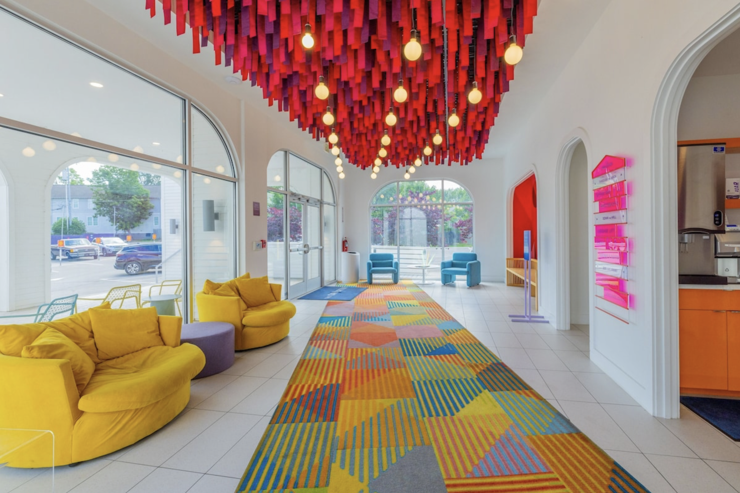 Bright hotel lobby with colorful decor including yellow couches, a vibrant patterned carpet, modern pink and red ceiling installation, blue chairs near large windows, and a reception area to the right.