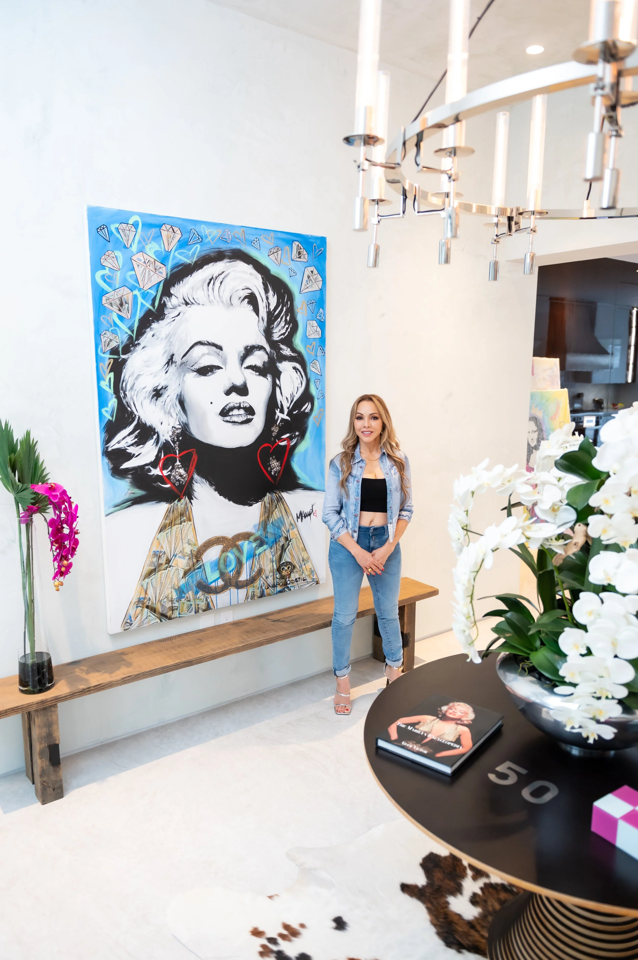 A woman standing in front of a large pop art portrait of Marilyn Monroe on a gallery wall, surrounded by flowers and modern decor.