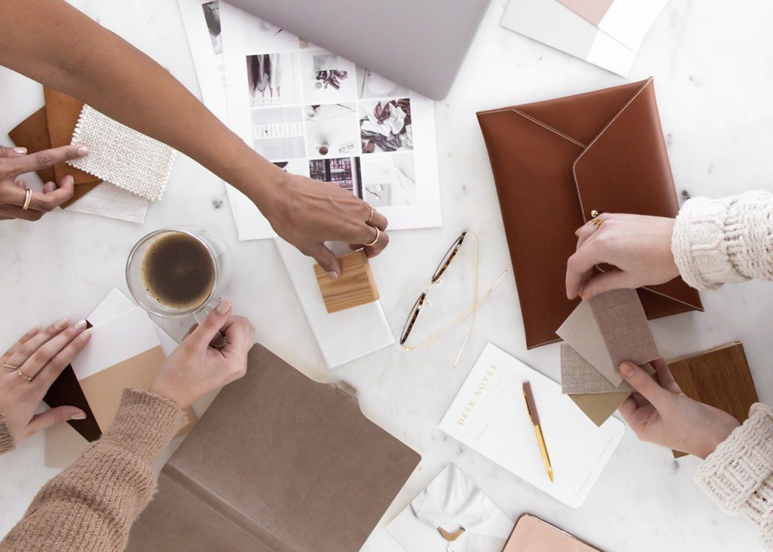 Overhead view of a workspace with two people working on a project, with notebooks, color swatches, glasses, a cup of coffee, and printed images on a white table.