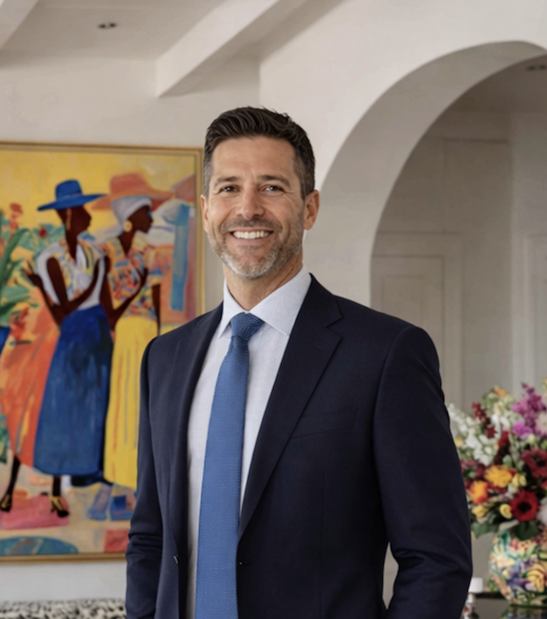 Man in a dark suit, white shirt, and blue tie standing indoors with colorful artwork and flowers behind him.