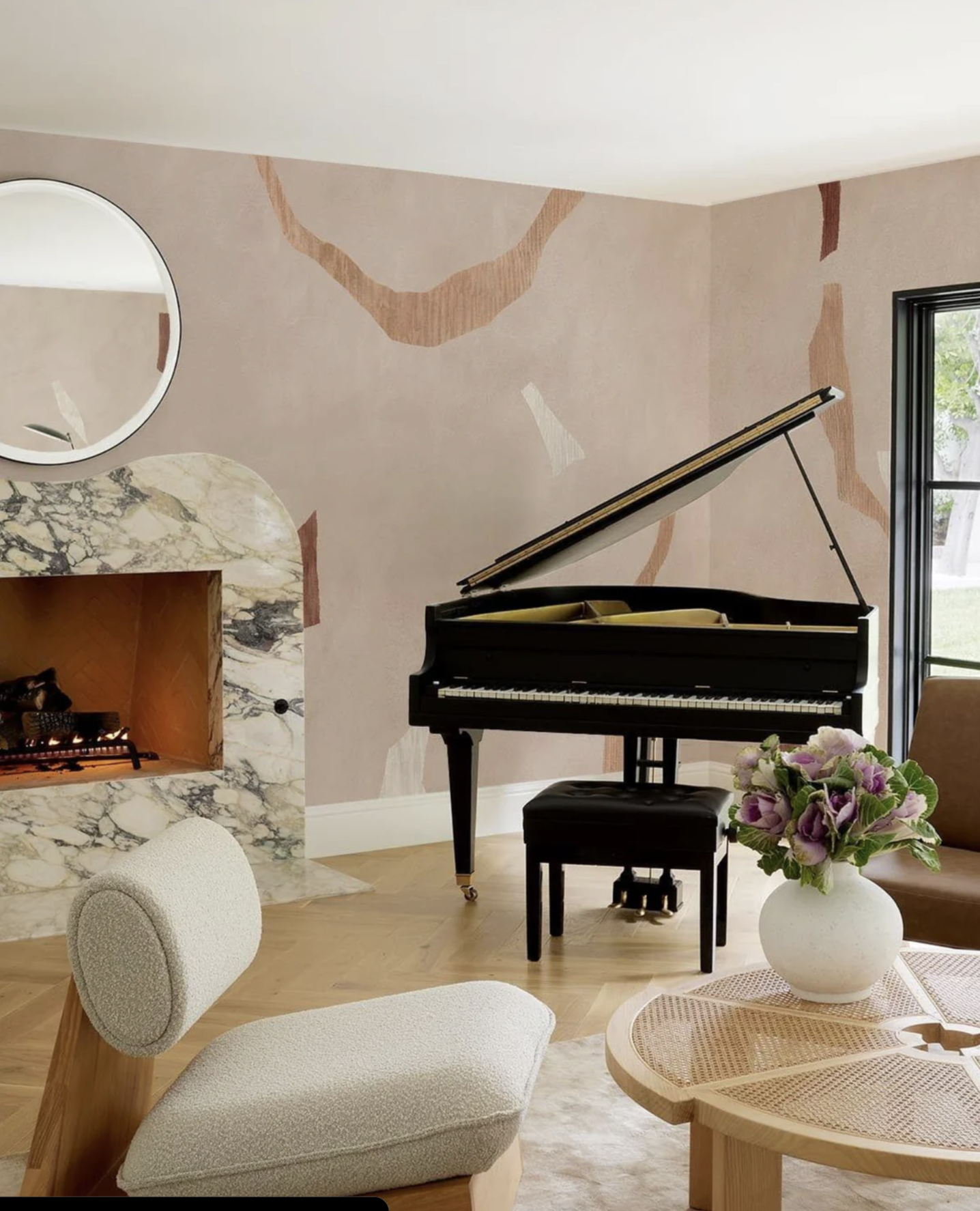 A living room with a grand piano, a corner fireplace with a marble surround, a round mirror above it, a cream upholstered chair, a wooden coffee table with a vase of pink and purple flowers, and a large window.