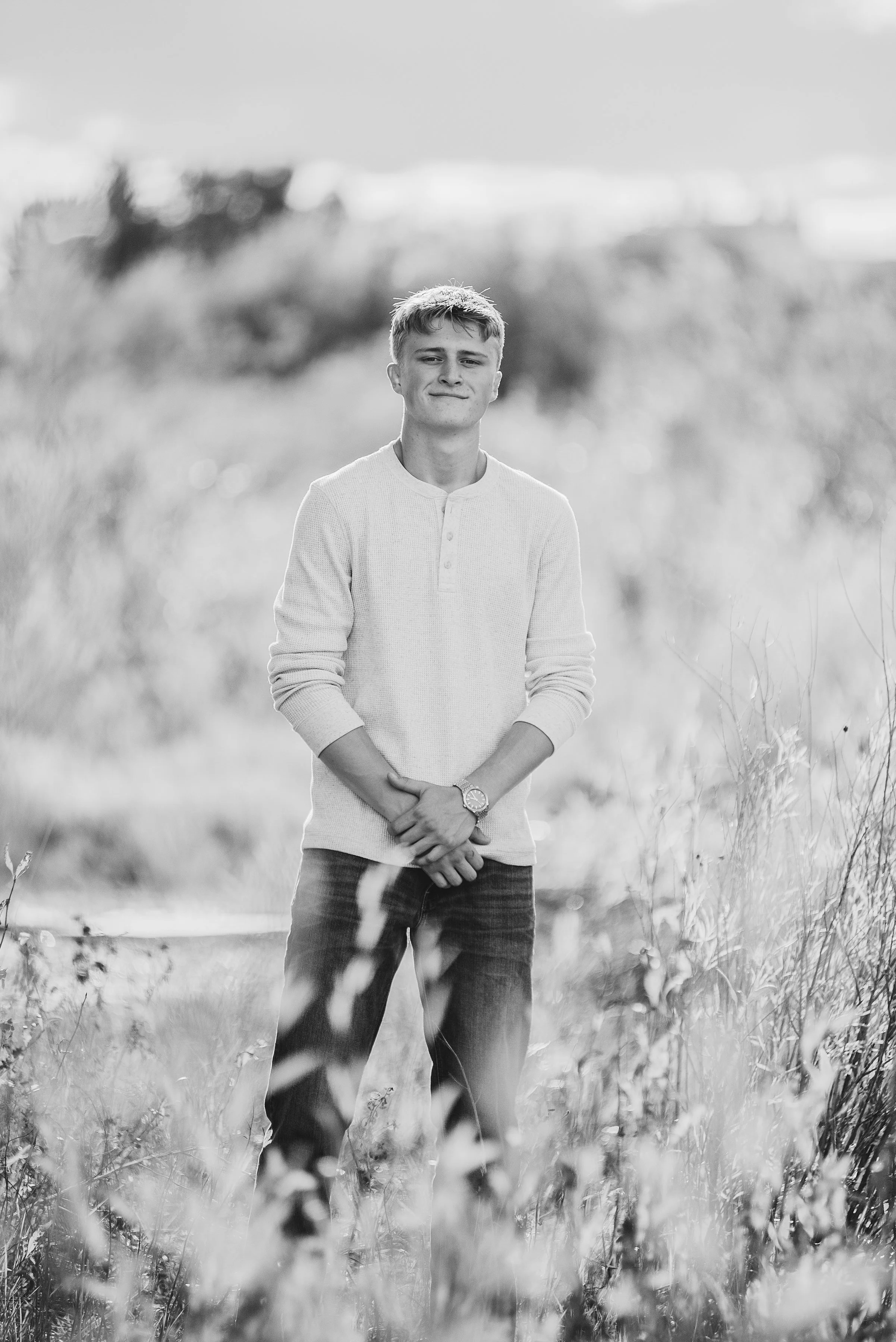 A young man standing outdoors in a field with tall grass, wearing a long-sleeve shirt and jeans, smiling softly at the camera in black and white.