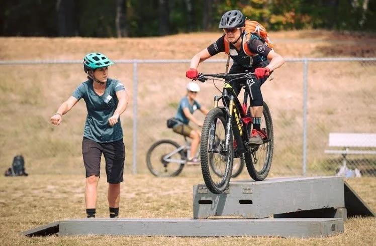 Bicycle rider jumping off a raised platform in an outdoor park with another person walking nearby and a person riding a bike in the background.