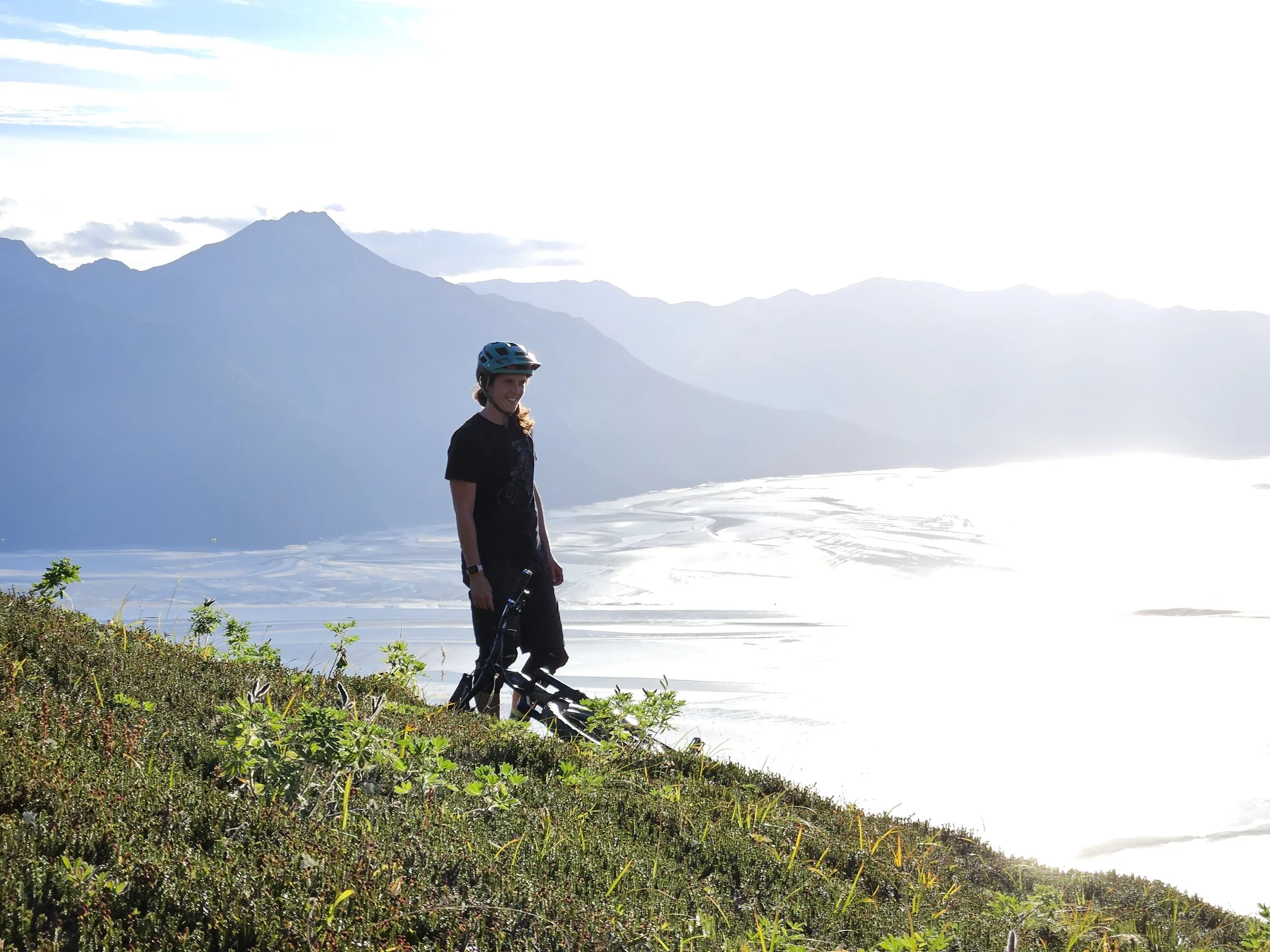 A person wearing a helmet smiling while standing outdoors at the base of a grassy hill with flowering plants, overlooking a large body of water with mountain peaks in the background on a sunny day.