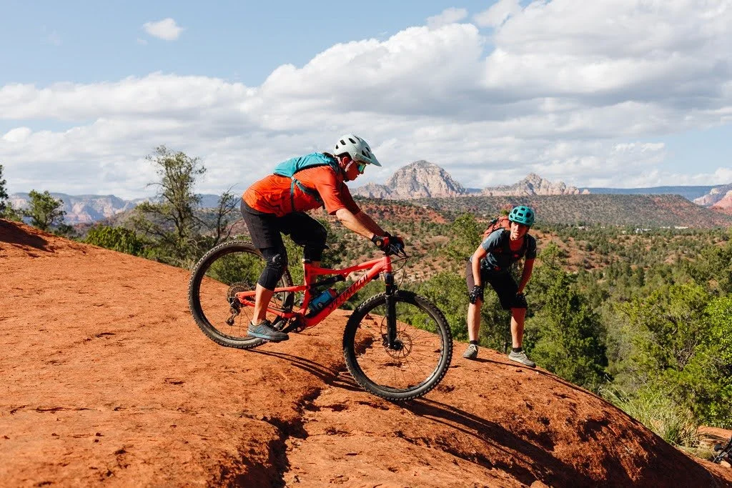 Two mountain bikers riding on a red dirt trail in a desert landscape with green shrubs, trees, and distant rocky mountains under a partly cloudy sky.