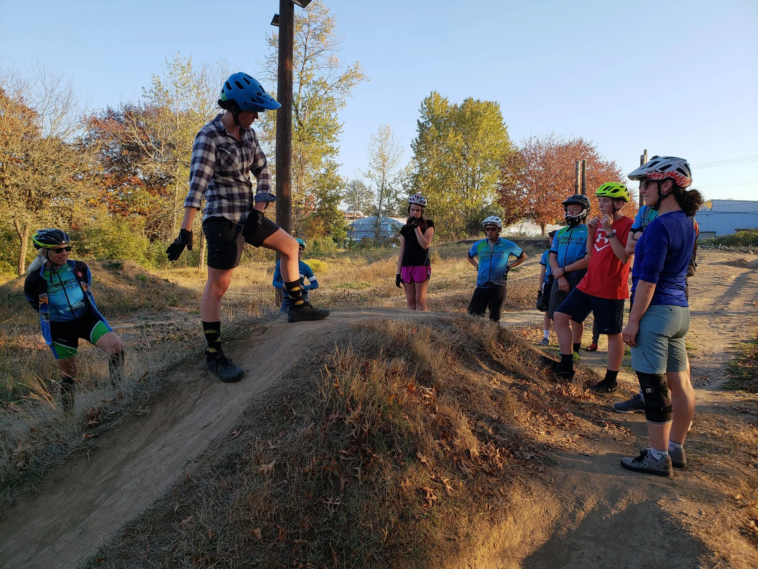 A group of people, mostly women, wearing helmets and athletic clothing, standing outdoors on a dirt trail with a slight incline, surrounded by trees with autumn foliage. A woman appears to be explaining or demonstrating something to the group.